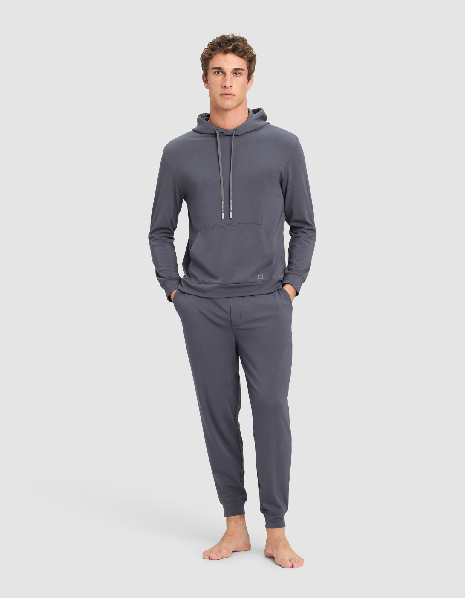 A young man stands barefoot against a plain background, hands in pockets, wearing the Cozy Earth Men's Brushed Bamboo Hoodie & Jogger Set in dark gray, and looks directly at the camera.