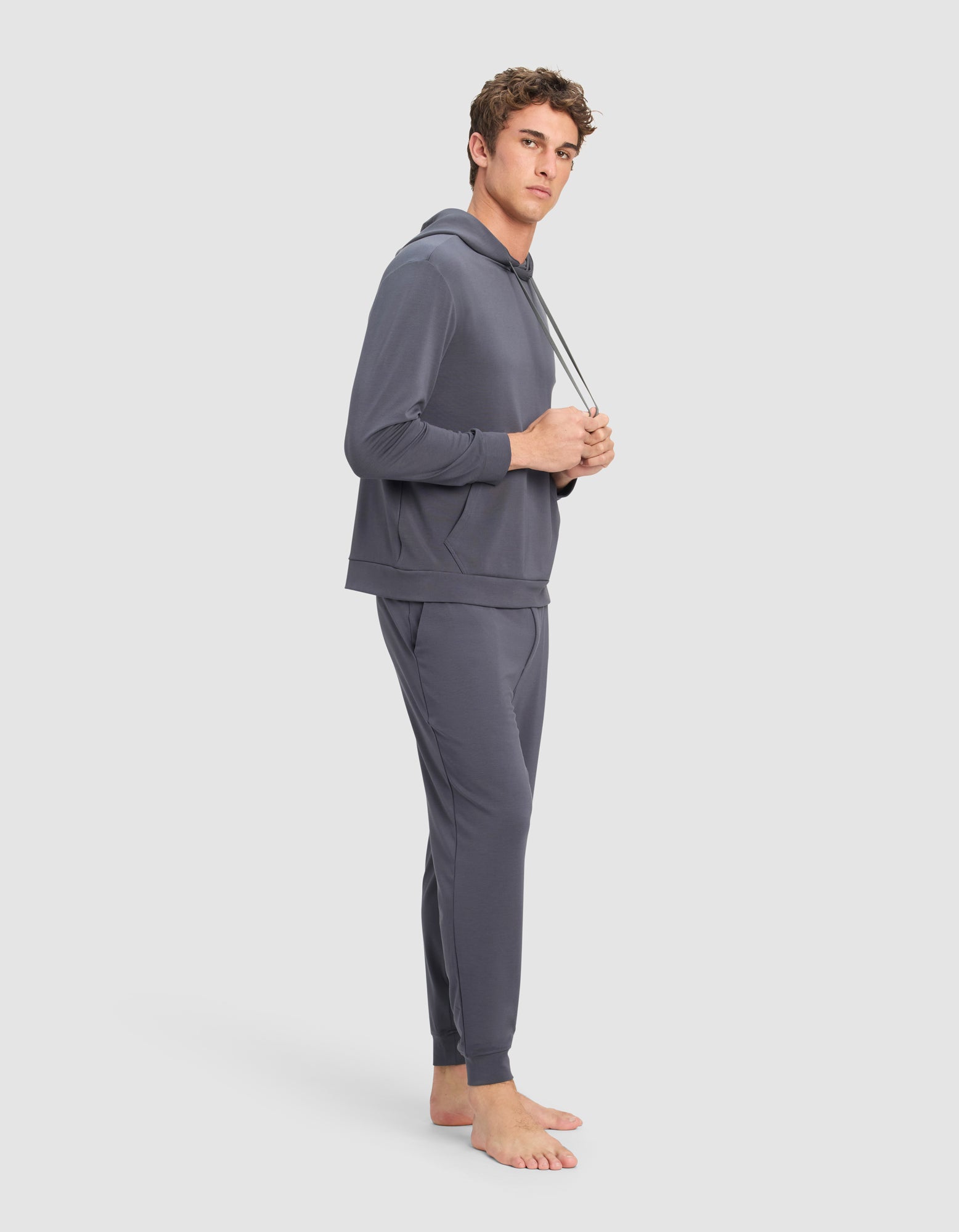 A young man stands barefoot against a plain light background, holding the drawstrings and looking at the camera, wearing the Cozy Earth Men's Brushed Bamboo Hoodie & Jogger Set in gray.