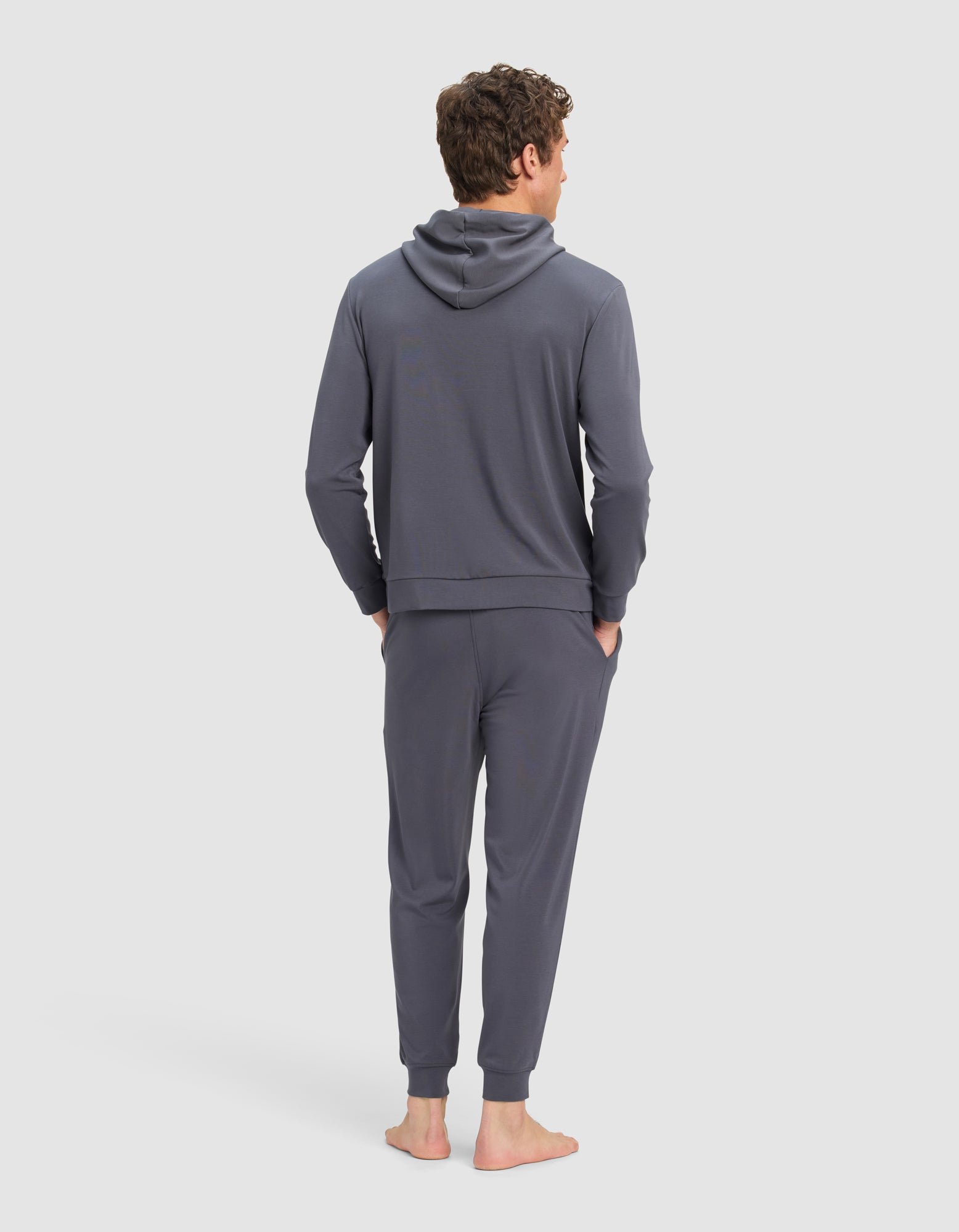 A man stands barefoot, facing away, wearing the Cozy Earth Men's Brushed Bamboo Hoodie & Jogger Set in dark gray with his hands in his pockets against a plain light background.