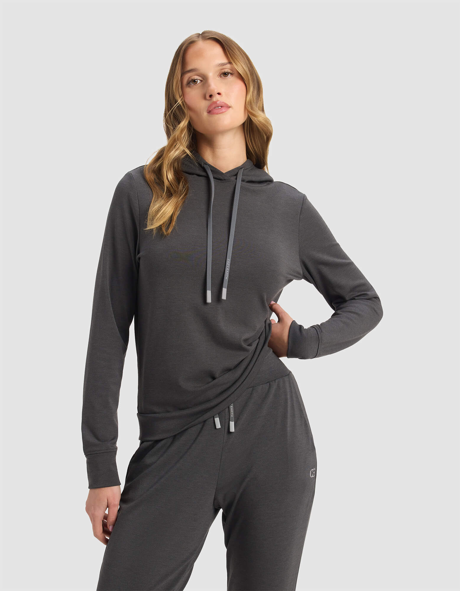 A woman with wavy blonde hair models the Cozy Earth Women's Brushed Bamboo Hoodie in charcoal, standing against a plain light gray background with one hand on her hip.