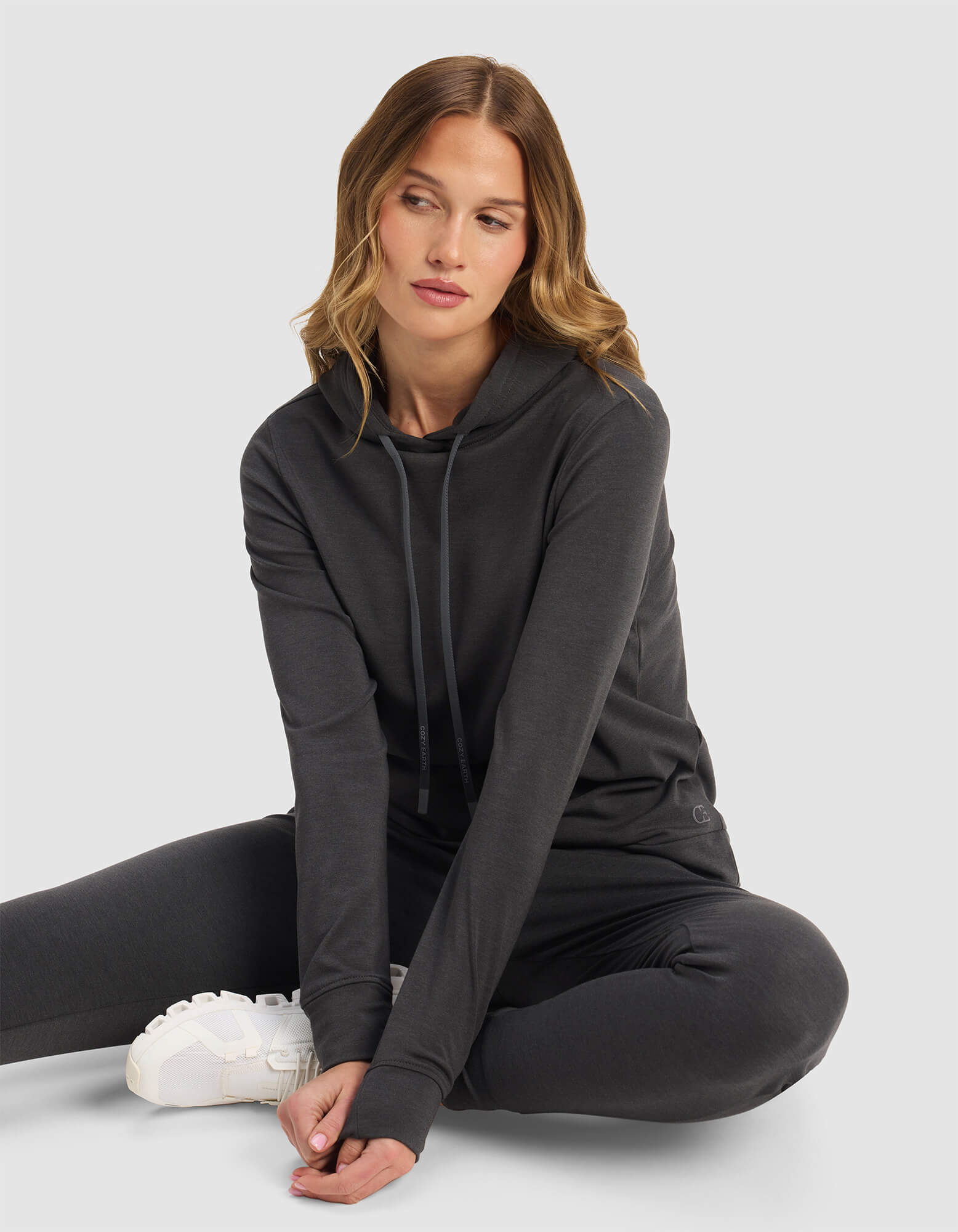 A woman with wavy, light brown hair sits on the floor in a relaxed pose, wearing Cozy Earth's Women's Brushed Bamboo Hoodie in dark gray with matching pants and white sneakers, set against a plain light gray background.