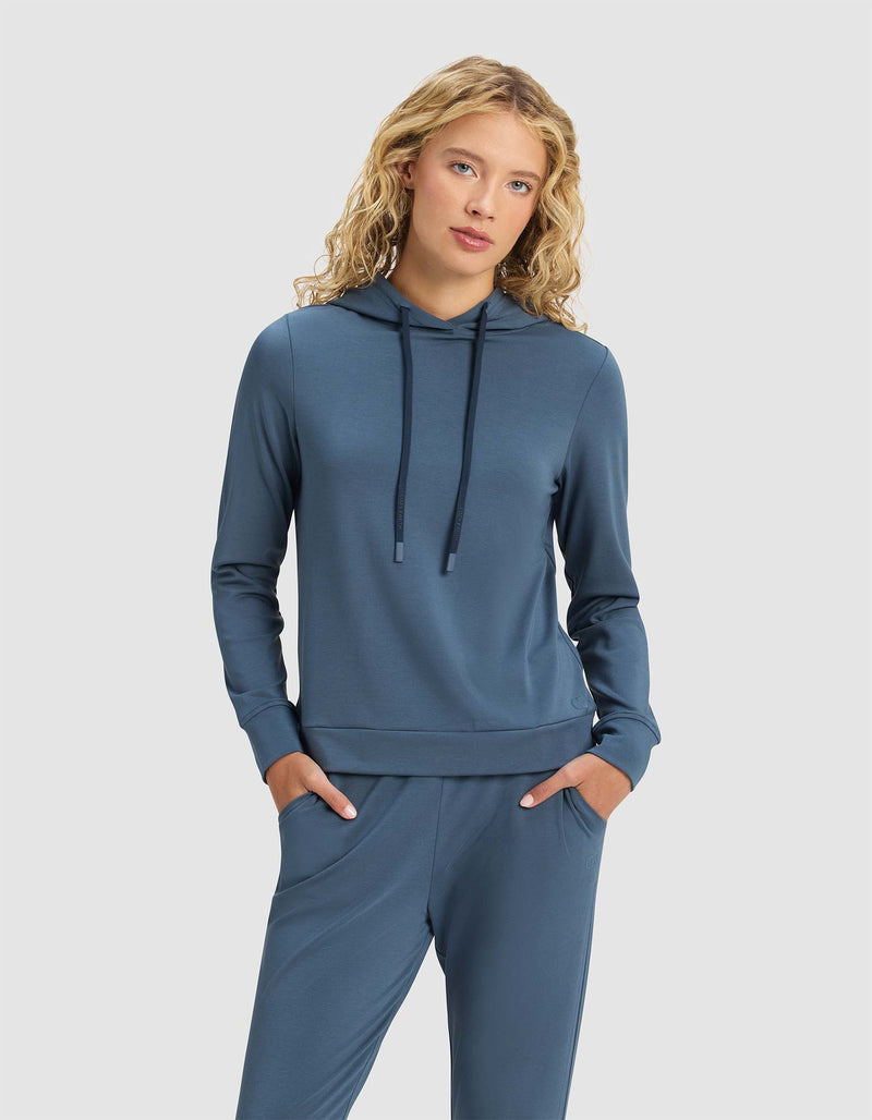 A woman with blonde curly hair models the Cozy Earth Women's Brushed Bamboo Hoodie in blue, paired with matching pants, standing hands-in-pockets against a plain light gray background.