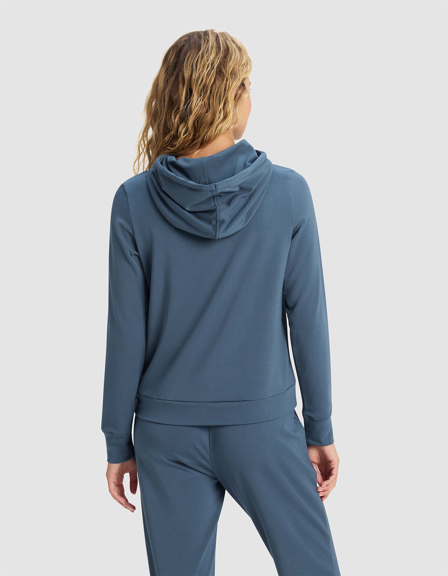 A woman with wavy, light brown hair is shown from the back wearing the Cozy Earth Women's Brushed Bamboo Hoodie in blue, with matching pants, against a plain light background. 
