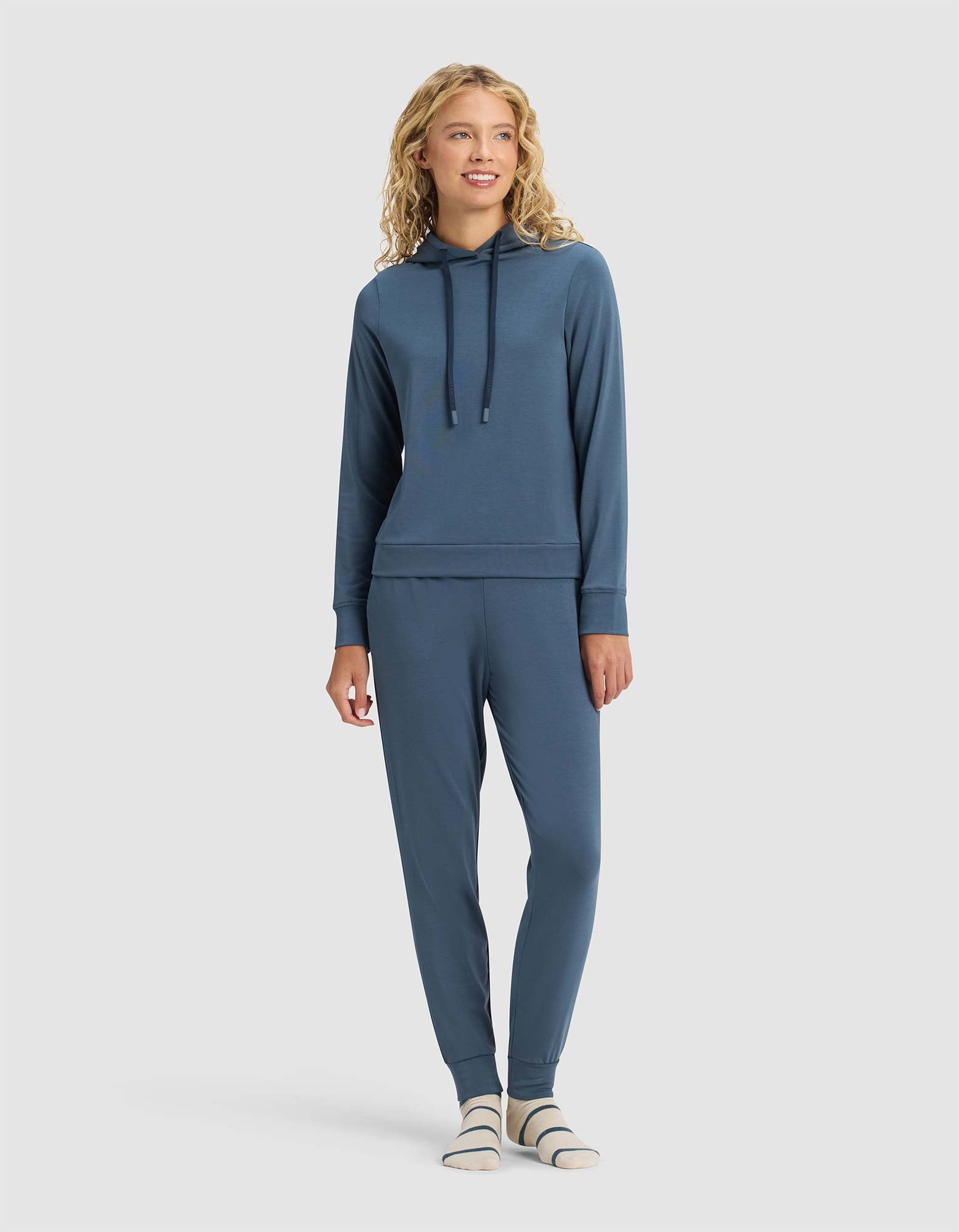 A woman with curly blonde hair stands and smiles against a plain light background, wearing Cozy Earth’s Women's Brushed Bamboo Hoodie in blue with matching jogger pants and white-gray striped socks. 