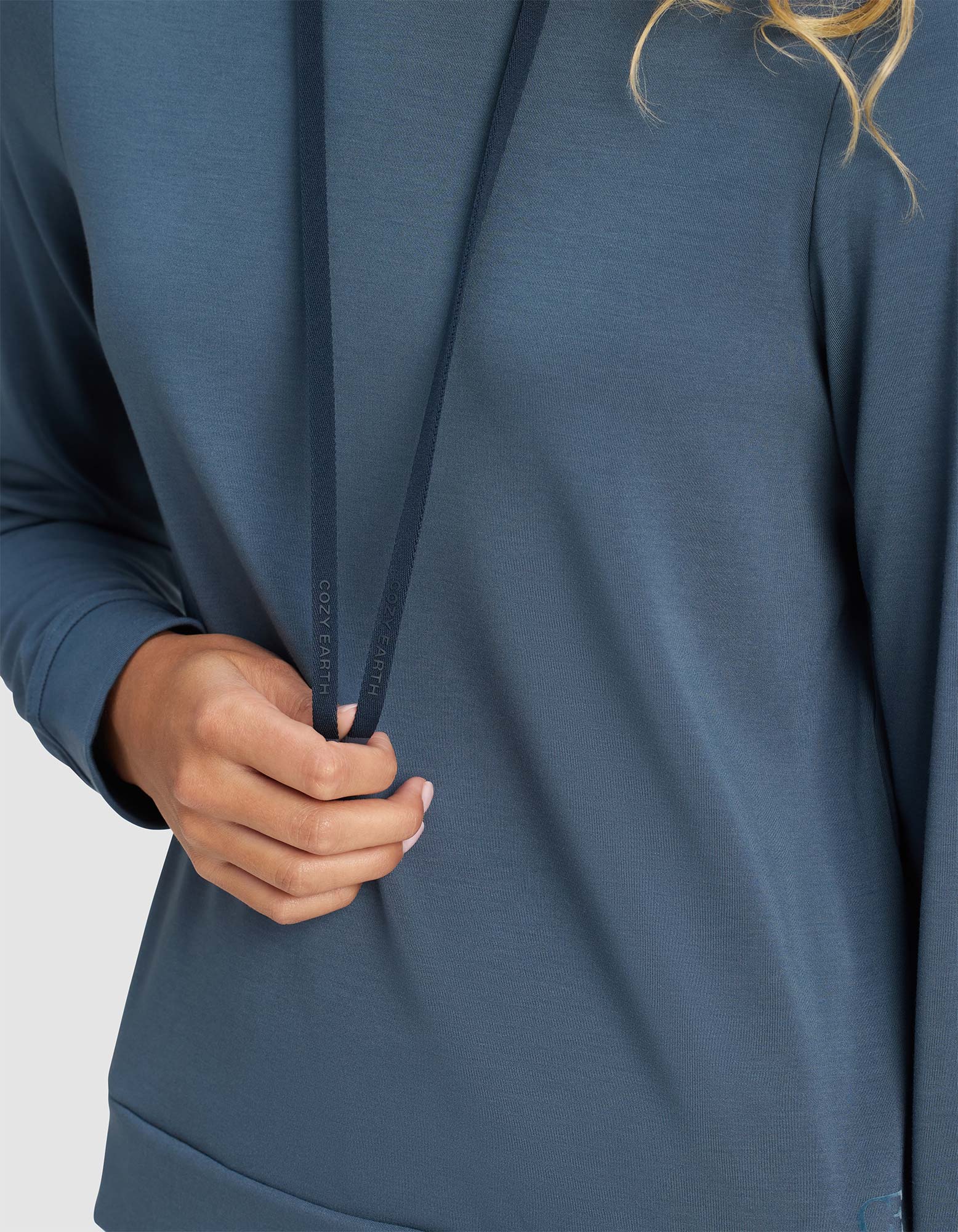 A person wears a Cozy Earth Women's Brushed Bamboo Hoodie in blue, holding one drawstring with their left hand. The visible part of the soft, comfortable hoodie highlights its cozy texture.