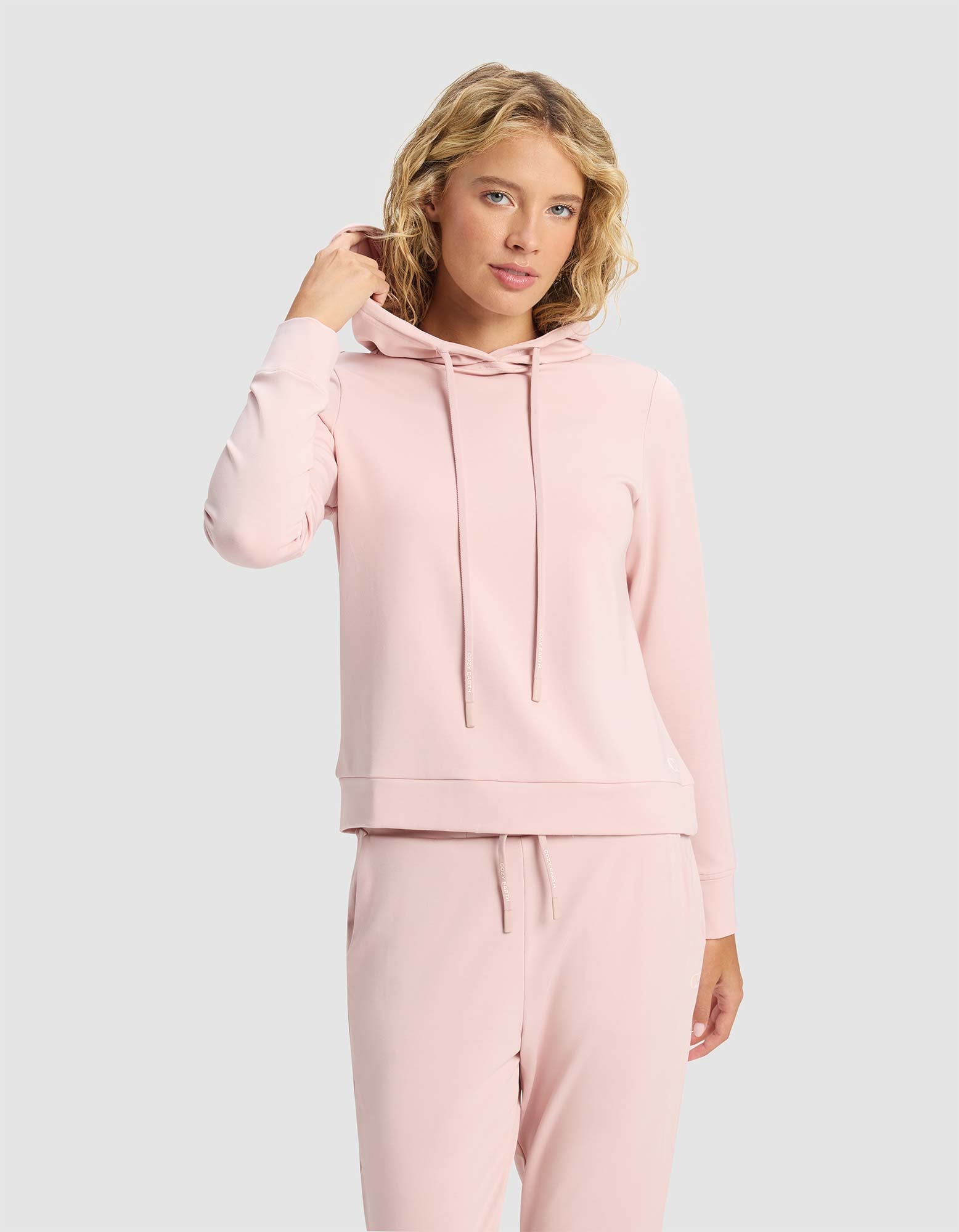 A woman with blond curly hair wears the Cozy Earth Women's Brushed Bamboo Hoodie in light pink, paired with matching sweatpants, against a plain light gray background. She faces the camera and touches her hair with one hand.