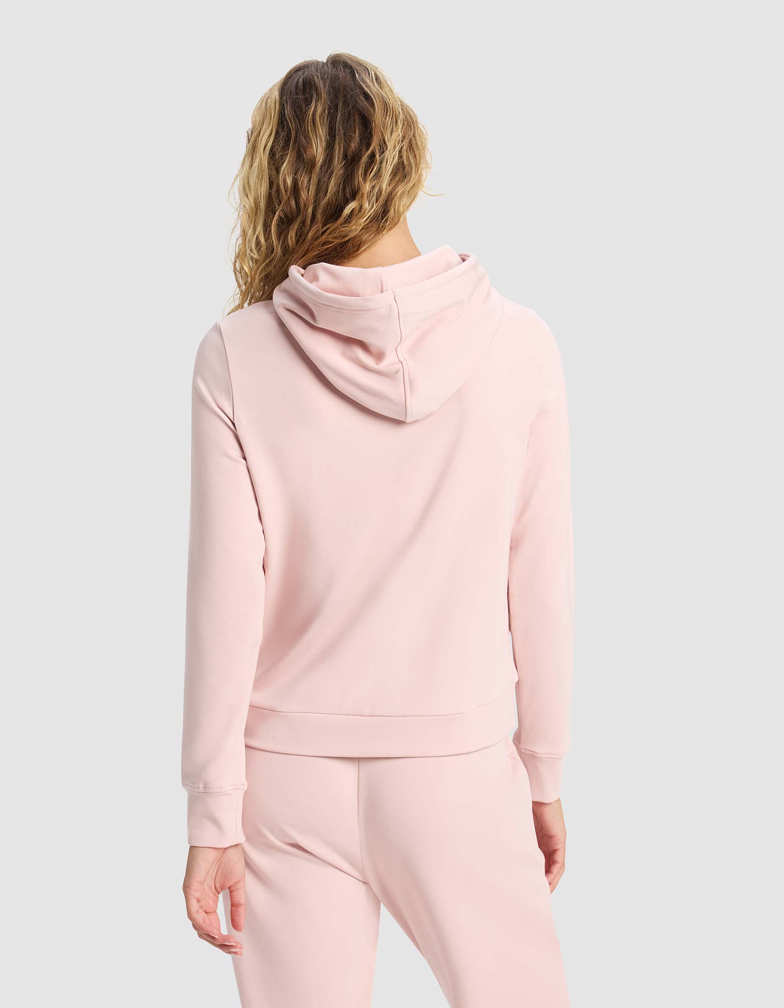 A woman with wavy blonde hair is seen from the back, wearing the Cozy Earth Women's Brushed Bamboo Hoodie in light pink, paired with matching pants, against a plain light gray background. 