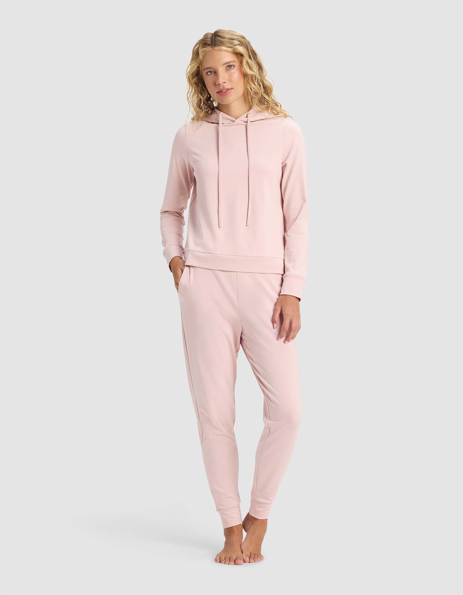 A woman with wavy blonde hair stands barefoot against a light gray background, facing forward. She wears a light pink Cozy Earth Women's Brushed Bamboo Hoodie and matching jogger pants, with one hand in her pocket. 