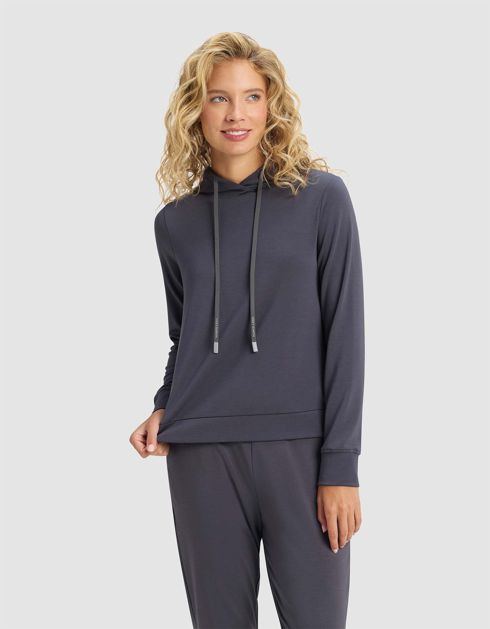 A woman with blonde, wavy hair stands against a light gray background, smiling softly while holding the hem of her Cozy Earth Women's Brushed Bamboo Hoodie, paired with matching pants.