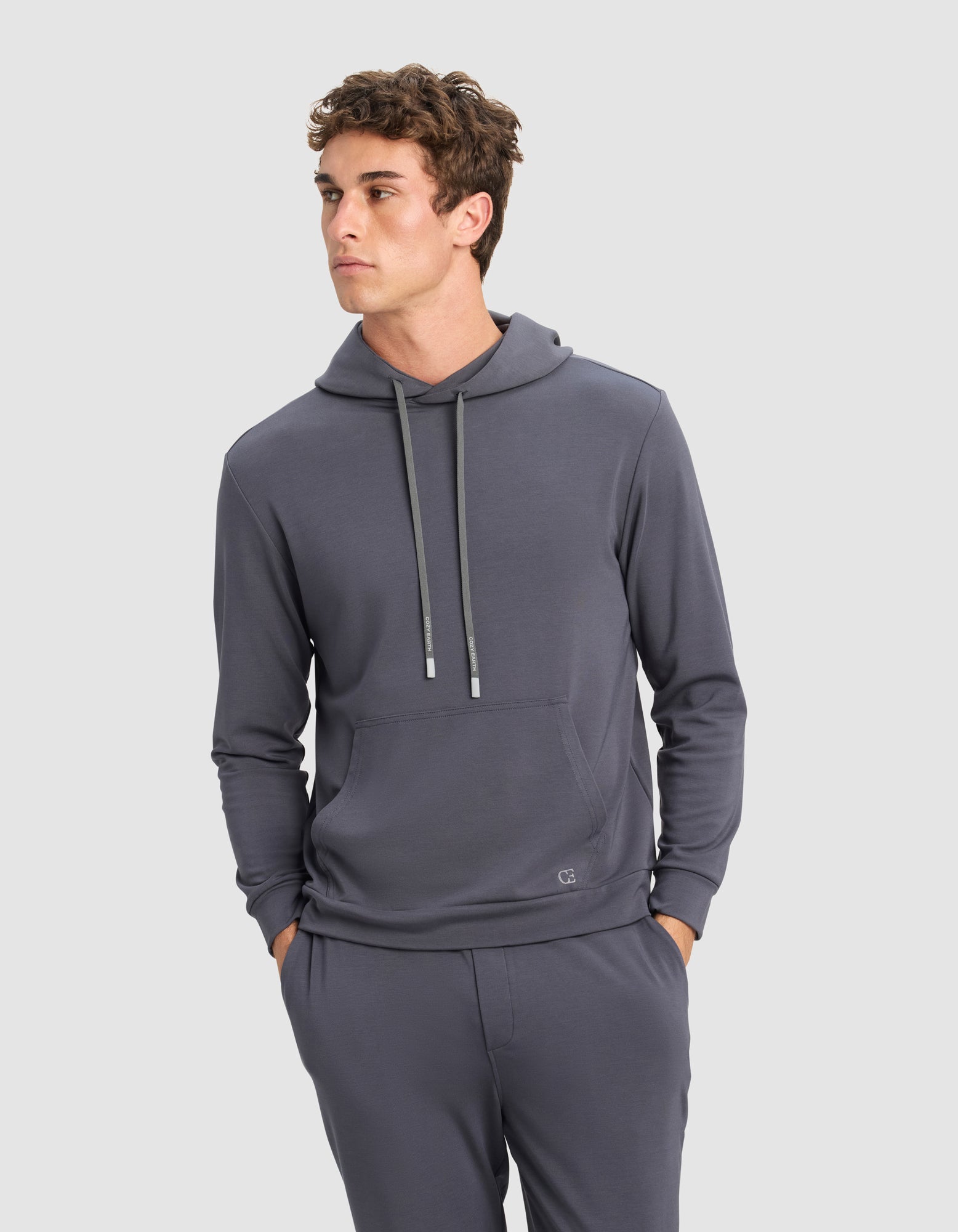 A young man models the Cozy Earth Men's Brushed Bamboo Hoodie & Jogger Set in gray, standing with his hands in his pockets and gazing slightly to the side against a plain light background.