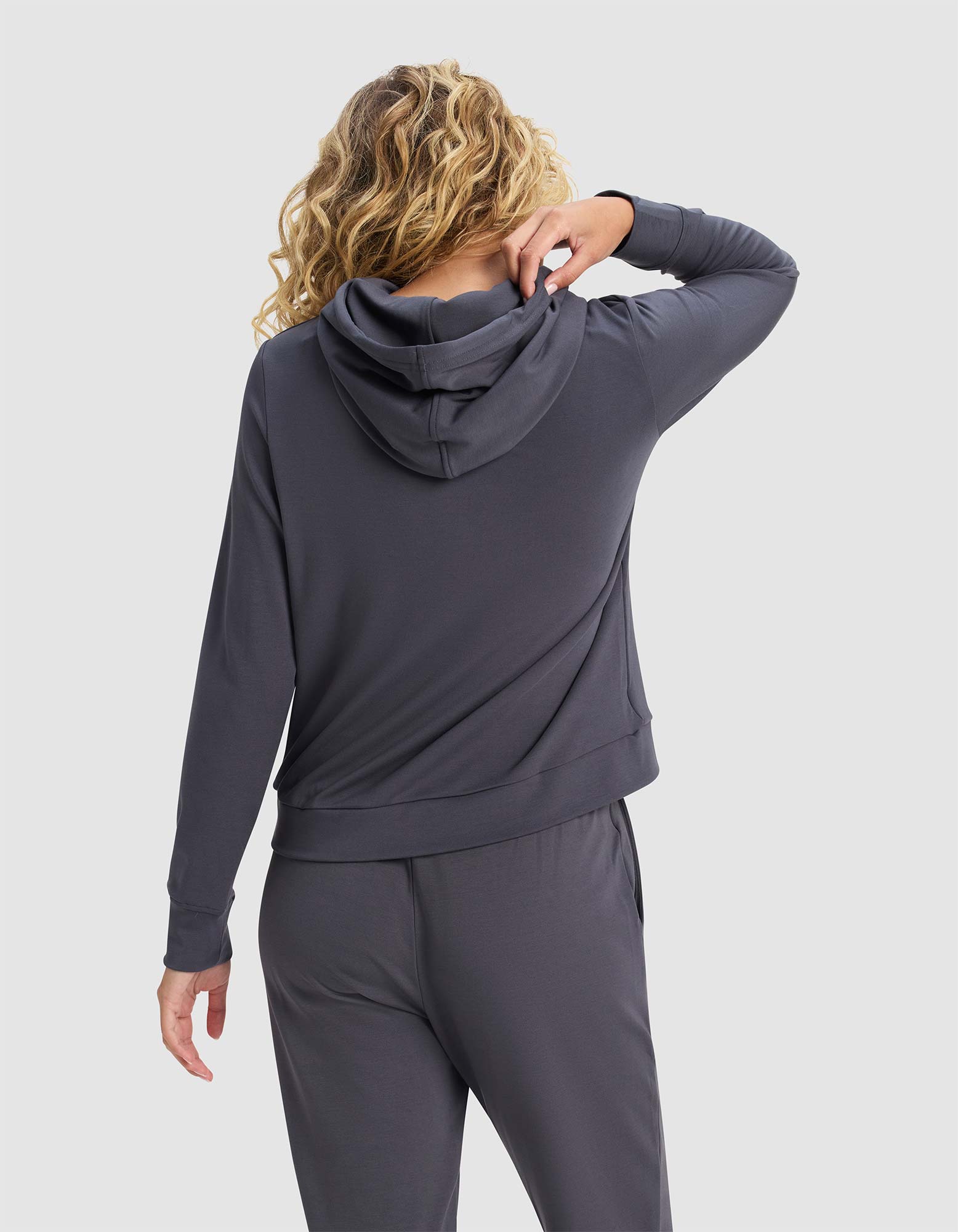 A woman with curly blonde hair, seen from the back, wears the Cozy Earth Women's Brushed Bamboo Hoodie in dark gray, paired with matching pants. She lifts one arm to touch her neck against a plain light gray background.