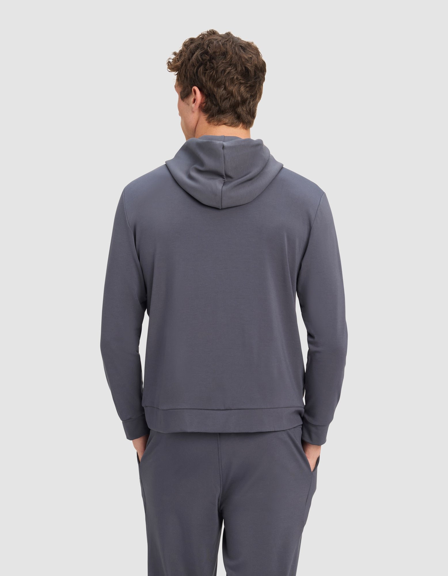 A man with curly brown hair stands with his back to the camera, hands in pockets, wearing the Cozy Earth Men's Brushed Bamboo Hoodie against a plain light background.