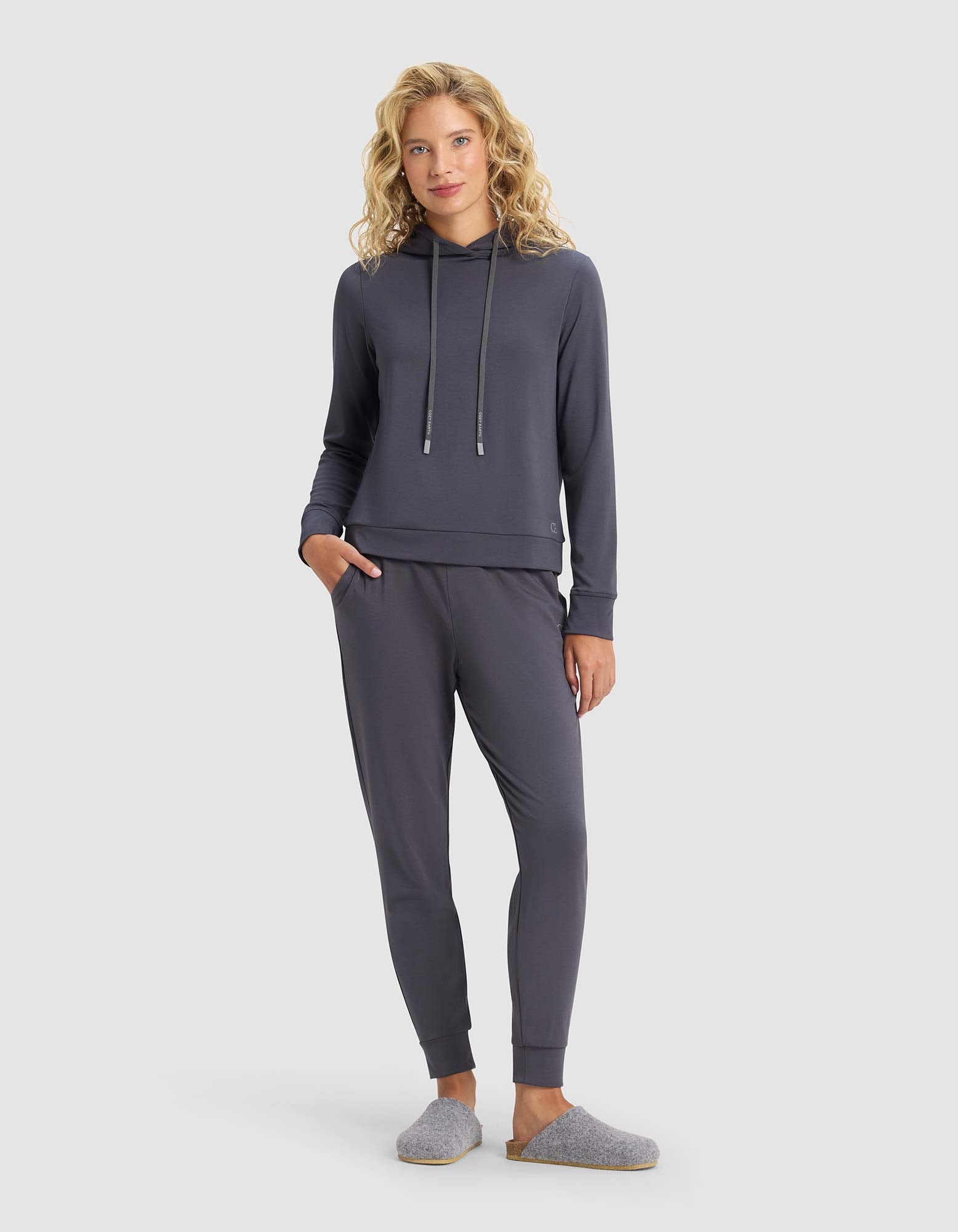A woman with curly blonde hair wears the Cozy Earth Women's Brushed Bamboo Hoodie in dark gray, paired with matching joggers and gray slippers, standing against a plain light background with her left hand in her pocket.