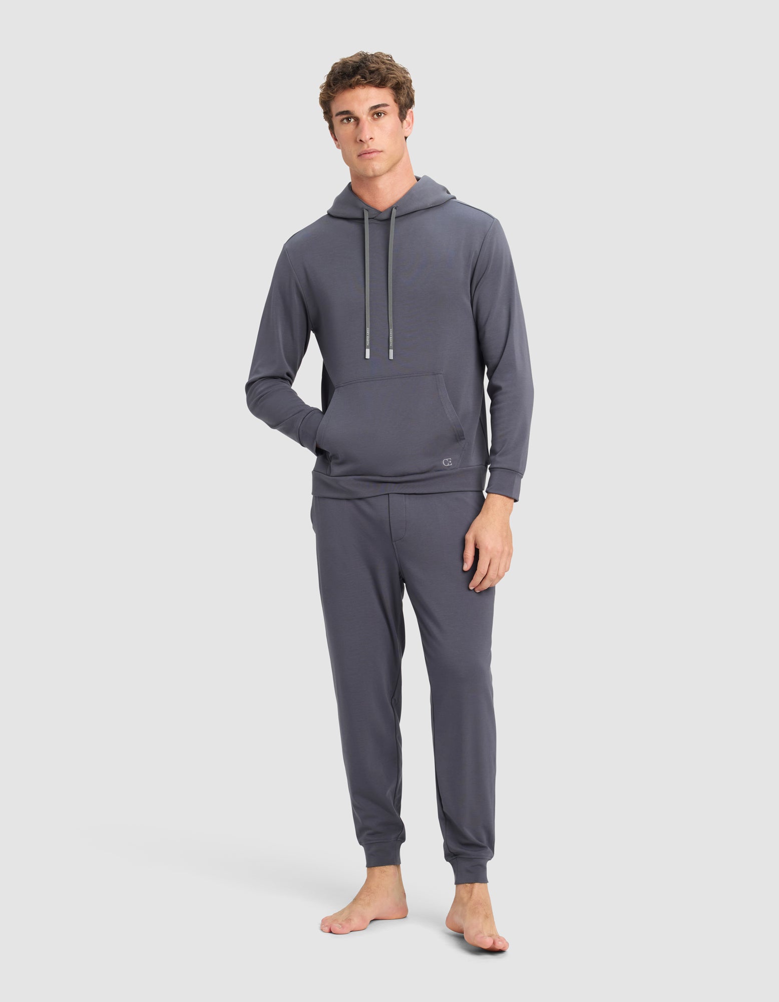 A young man stands barefoot on a light background, wearing the Cozy Earth Men's Brushed Bamboo Hoodie in dark gray with one hand in his pocket.