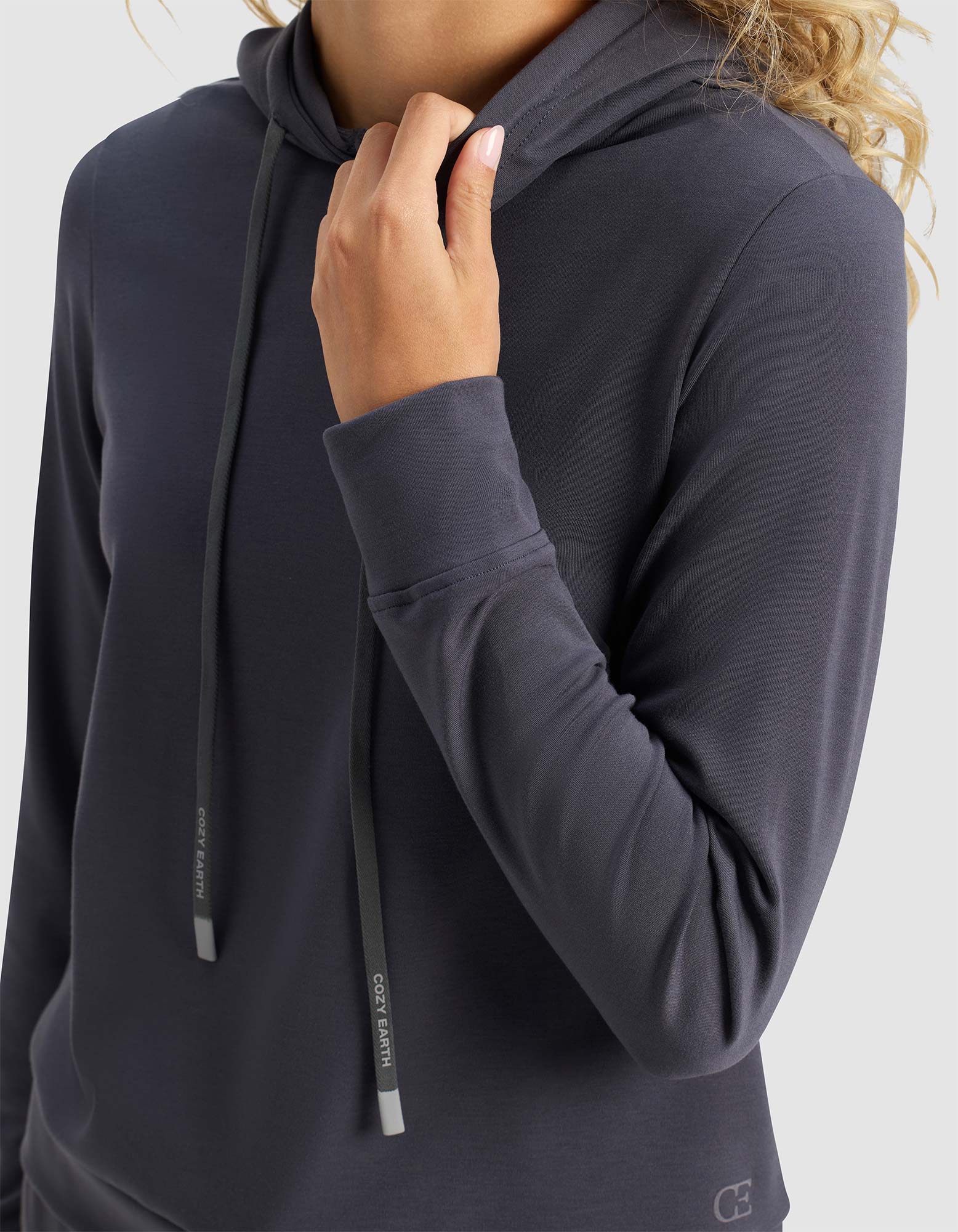 A person models the Cozy Earth Women's Brushed Bamboo Hoodie in dark gray, featuring long sleeves and a hood with drawstrings. They're holding the collar with one hand against a plain, light background; their face is not visible.