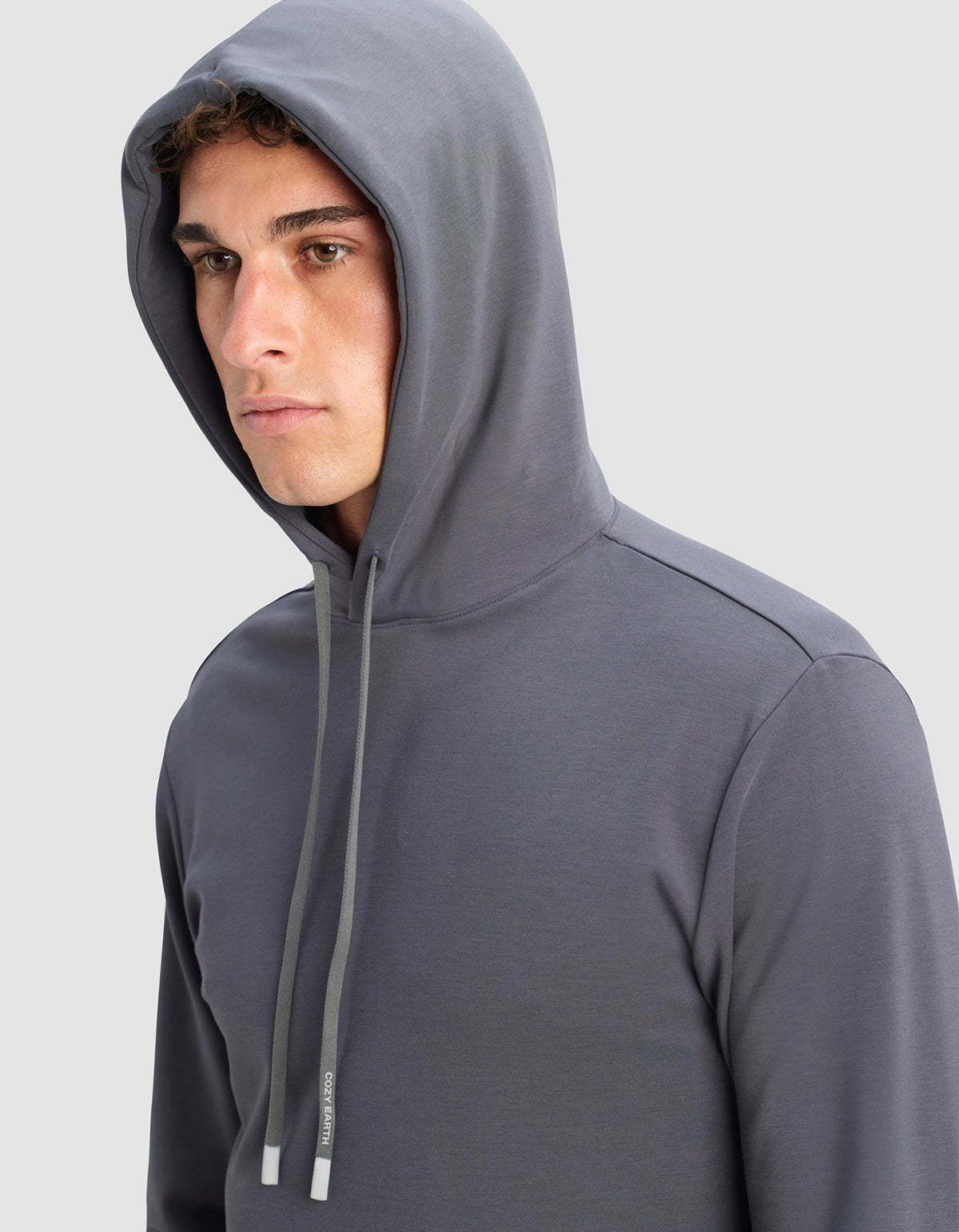 A young man wears the Cozy Earth Men's Brushed Bamboo Hoodie in gray with the hood up, looking slightly to the side against a light gray background.