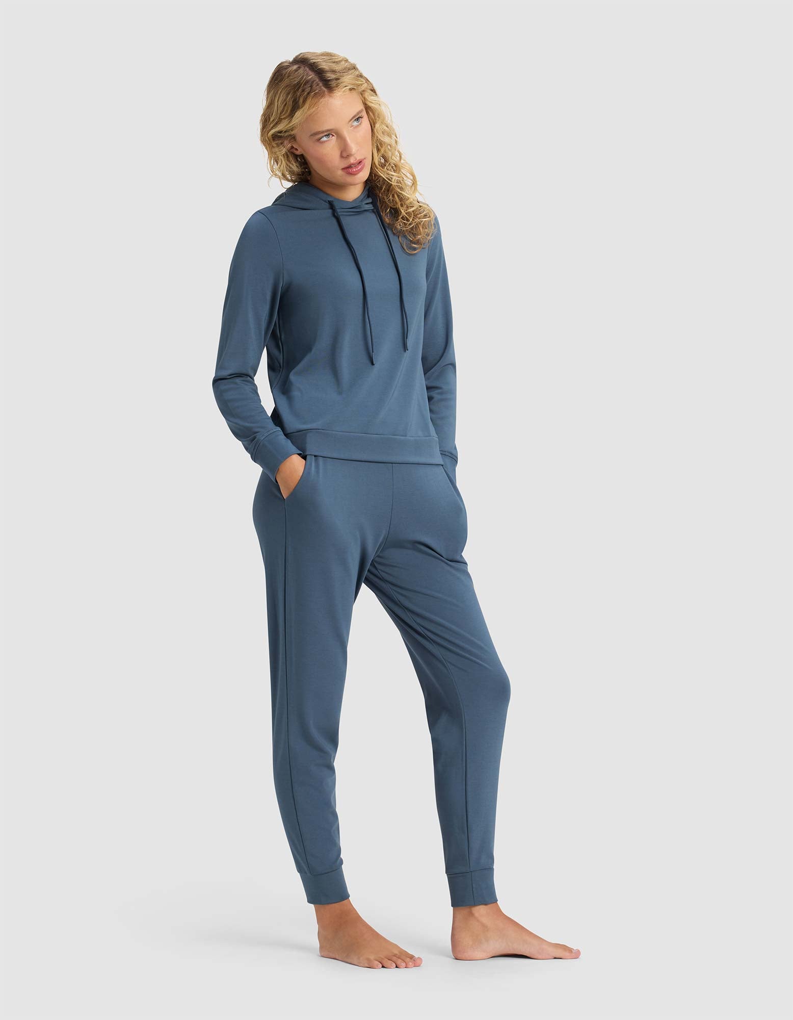 A woman with curly blonde hair models the Cozy Earth Women's Brushed Bamboo Hoodie & Jogger Set in blue, standing barefoot with hands in her pockets against a plain light background.|Color:Dawn