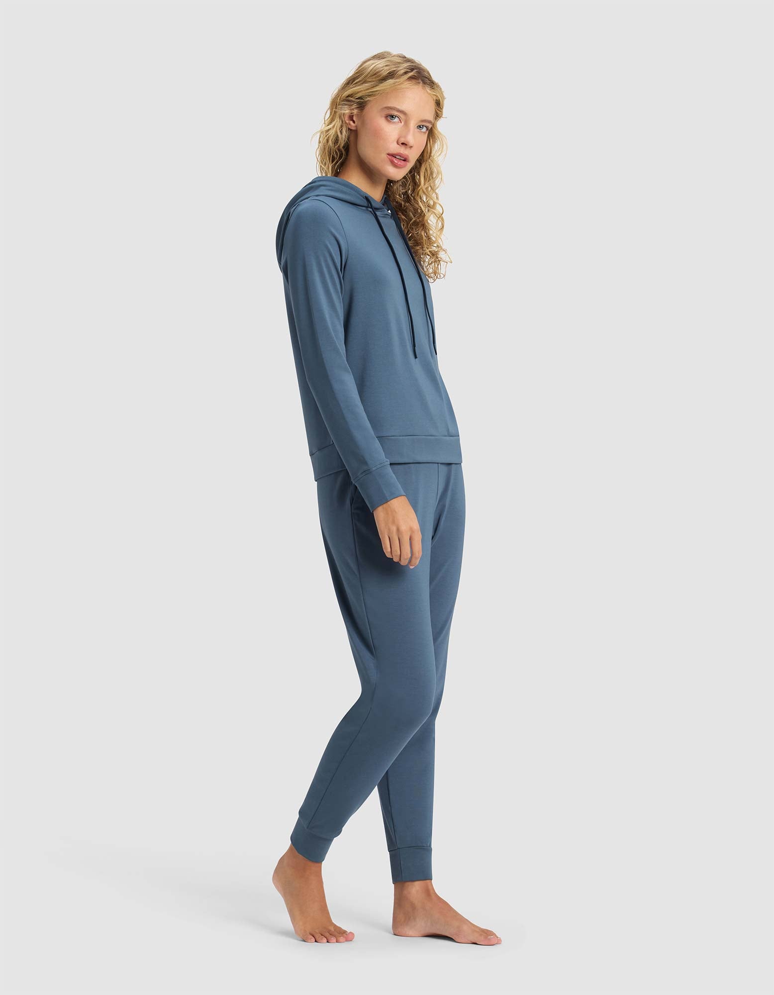 A woman with long curly blonde hair models the Cozy Earth Women's Brushed Bamboo Hoodie & Jogger Set, standing barefoot on a plain light background and glancing over her shoulder.