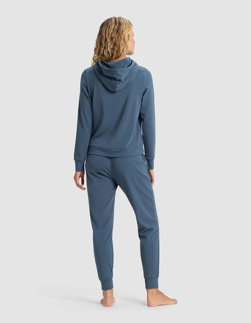 A woman with wavy blonde hair stands barefoot, facing away, wearing Cozy Earth's Women's Brushed Bamboo Hoodie & Jogger Set in blue against a plain light gray background.
