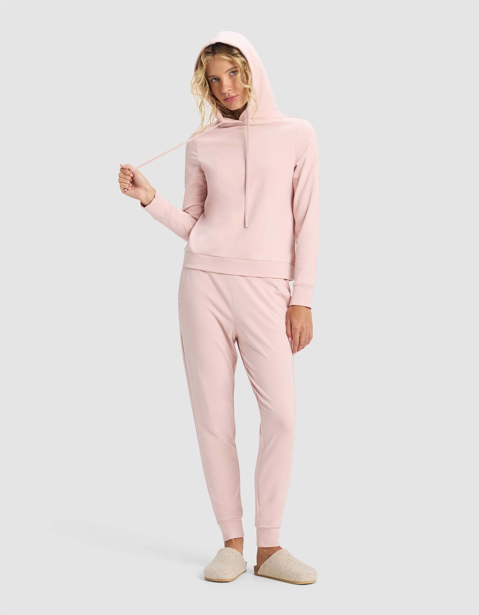 A woman wears the Cozy Earth Women's Brushed Bamboo Hoodie & Jogger Set in light pink with beige slippers, standing against a gray background and holding one drawstring of her hoodie.