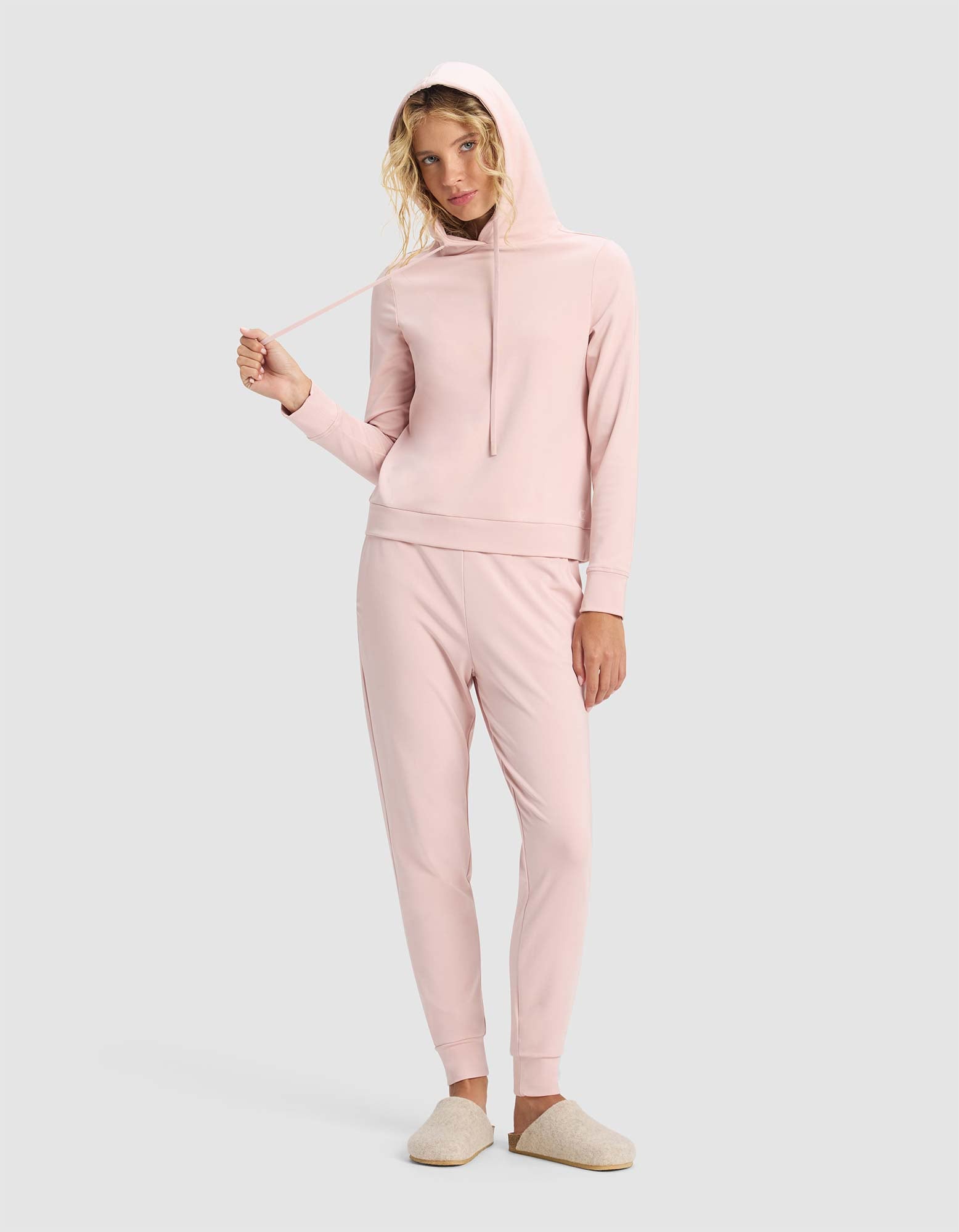 A woman wears the Cozy Earth Women's Brushed Bamboo Hoodie & Jogger Set in light pink with beige slippers, standing against a gray background and holding one drawstring of her hoodie.