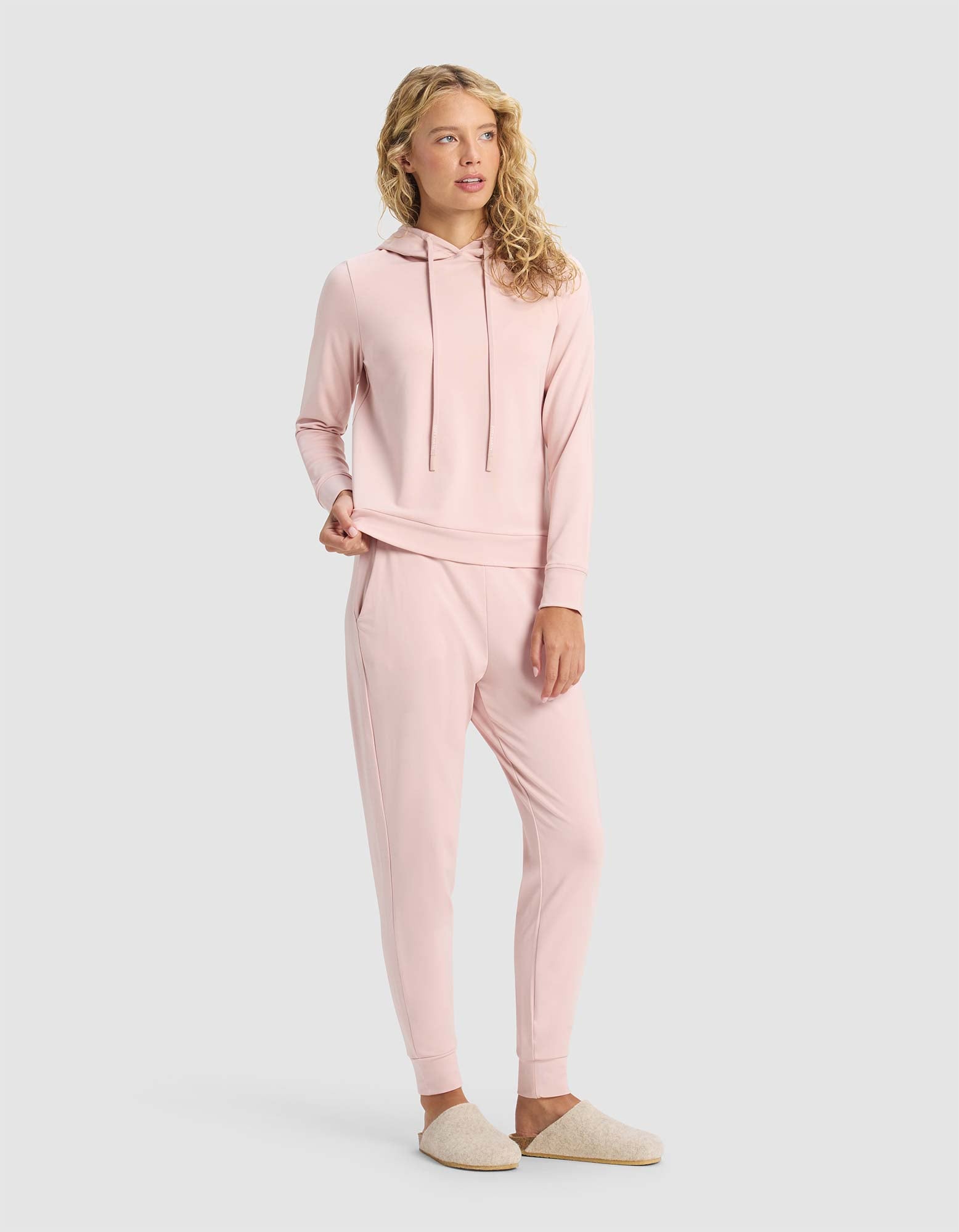 A woman wears the Cozy Earth Women's Brushed Bamboo Hoodie & Jogger Set in light pink with beige slippers. She has wavy blonde hair and looks to the side against a plain light gray background.