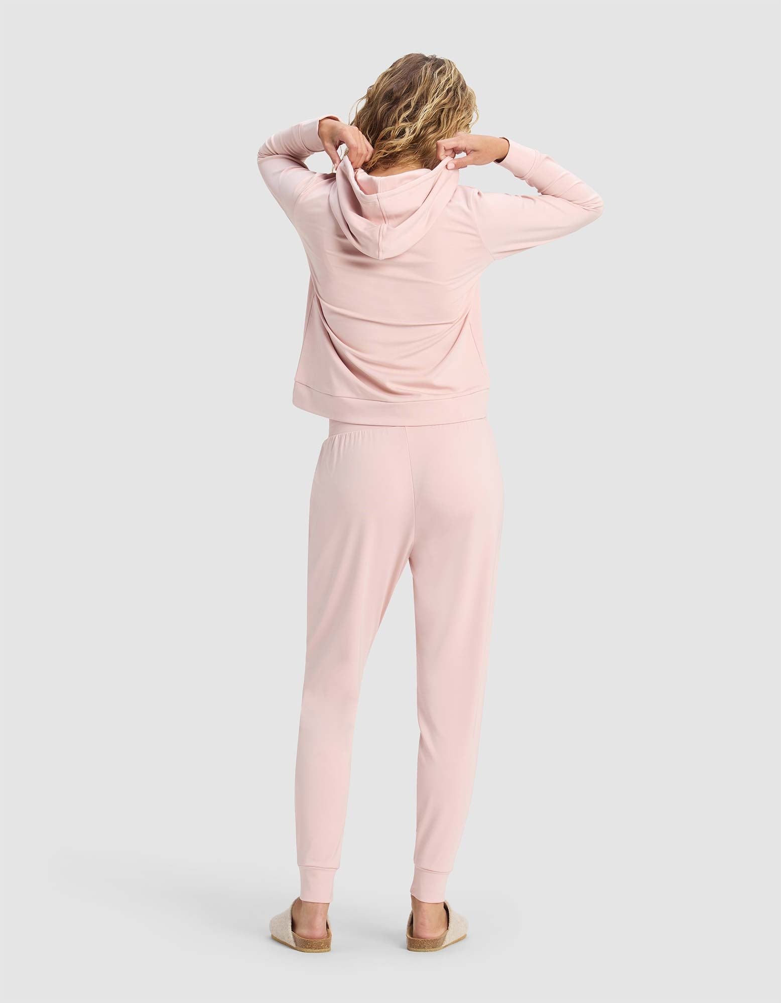 A person with wavy hair faces away, adjusting the hood of Cozy Earth's Women's Brushed Bamboo Hoodie & Jogger Set in light pink, paired with slippers, standing against a plain light gray background.