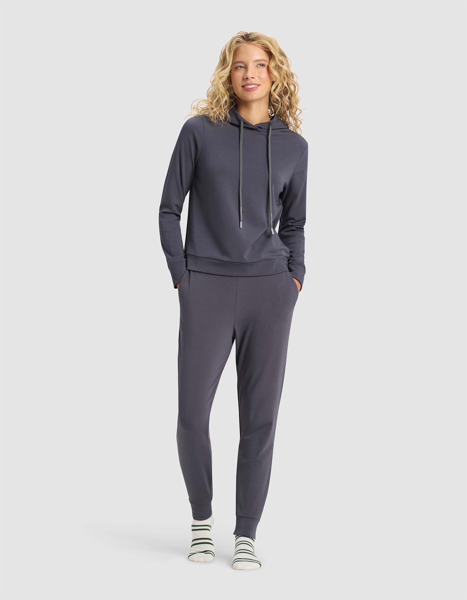 A woman with curly blonde hair smiles with hands in pockets, wearing Cozy Earth's Women's Brushed Bamboo Hoodie & Jogger Set in dark gray and white socks with green stripes.