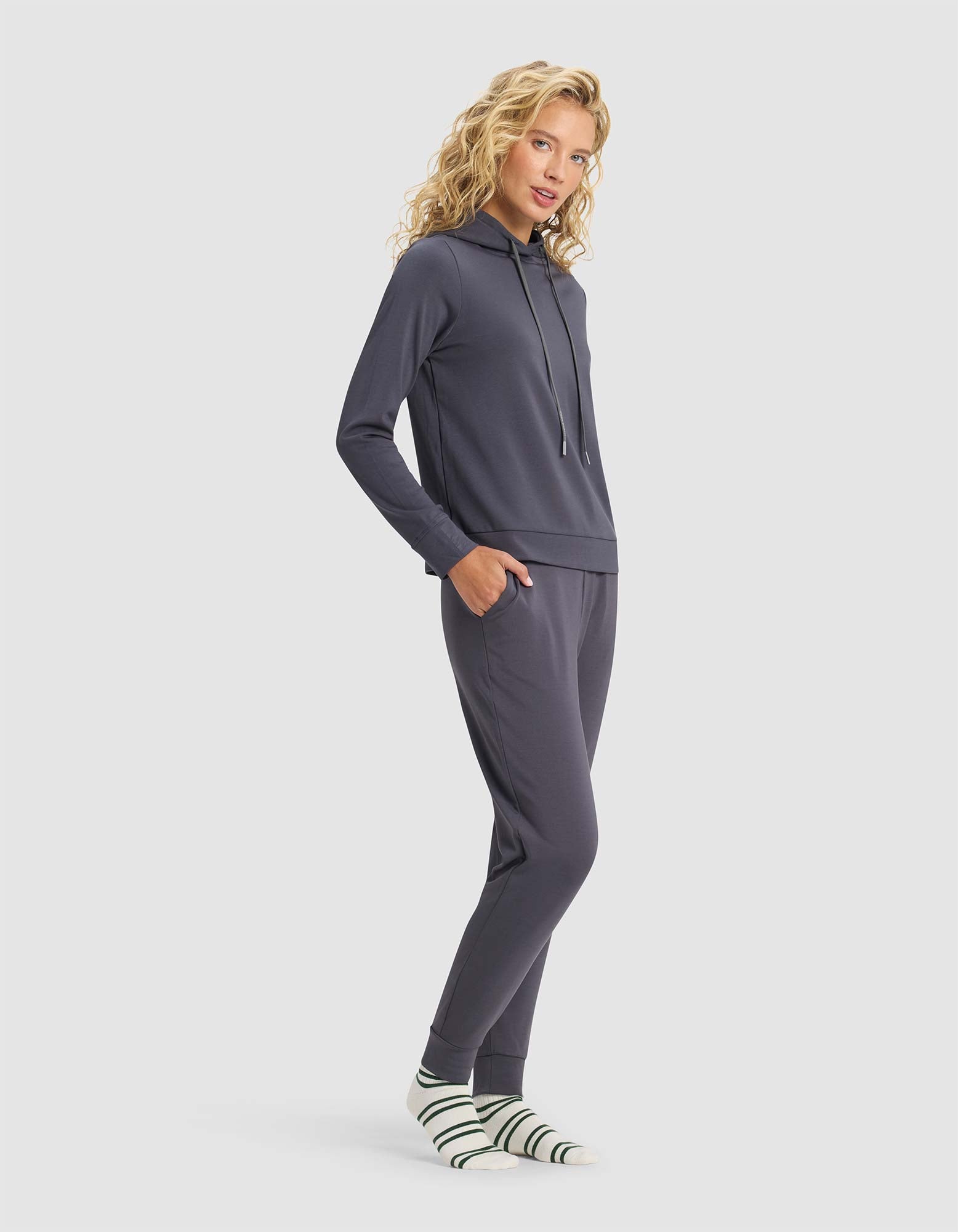 A woman with blonde curly hair models the Cozy Earth Women's Brushed Bamboo Hoodie & Jogger Set in dark gray, paired with white socks with green stripes. She stands sideways, hands in pockets, smiling against a light gray background.