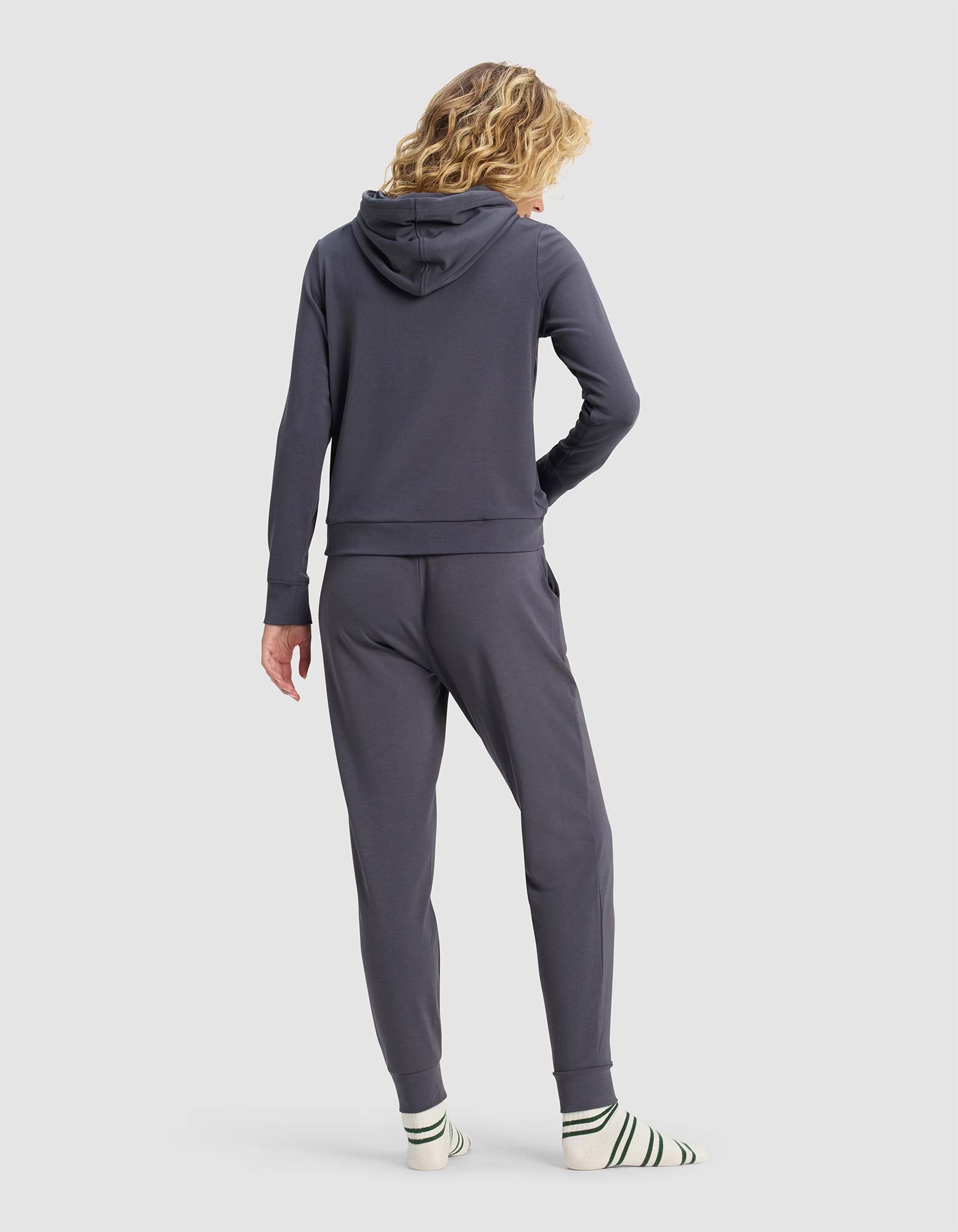 A woman with wavy blonde hair faces away, wearing the Cozy Earth Women's Brushed Bamboo Hoodie & Jogger Set in dark gray, paired with white socks featuring green and black stripes against a plain light gray background.