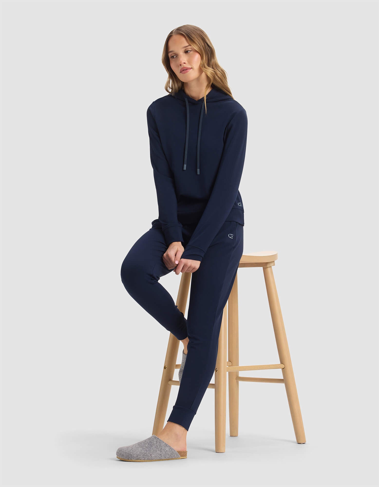A woman in the Women's Brushed Bamboo Hoodie by Cozy Earth and matching joggers sits on a wooden stool, one foot down. She has loose hair, gray slippers, and poses against a plain light background.