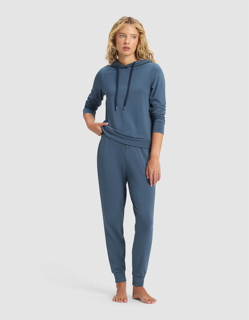A woman with wavy blonde hair wears Cozy Earth’s Women’s Brushed Bamboo Jogger Pant in blue with a matching hooded sweatshirt, standing barefoot against a plain light background.