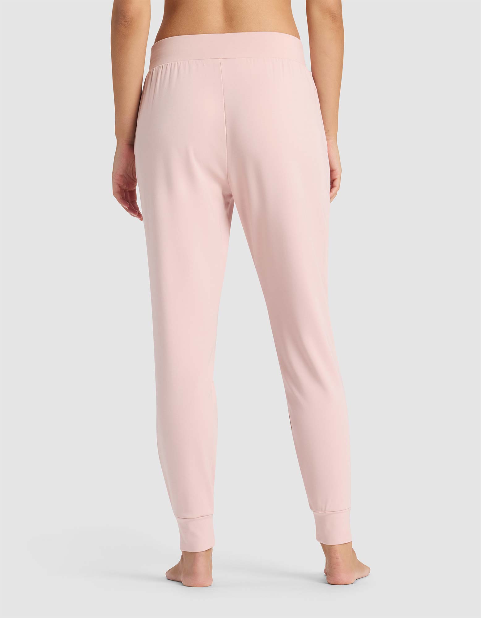 A person stands barefoot facing away, wearing Cozy Earth's Women's Brushed Bamboo Jogger Pant in light pink with an elastic waistband and fitted cuffs, set against a plain light gray background.