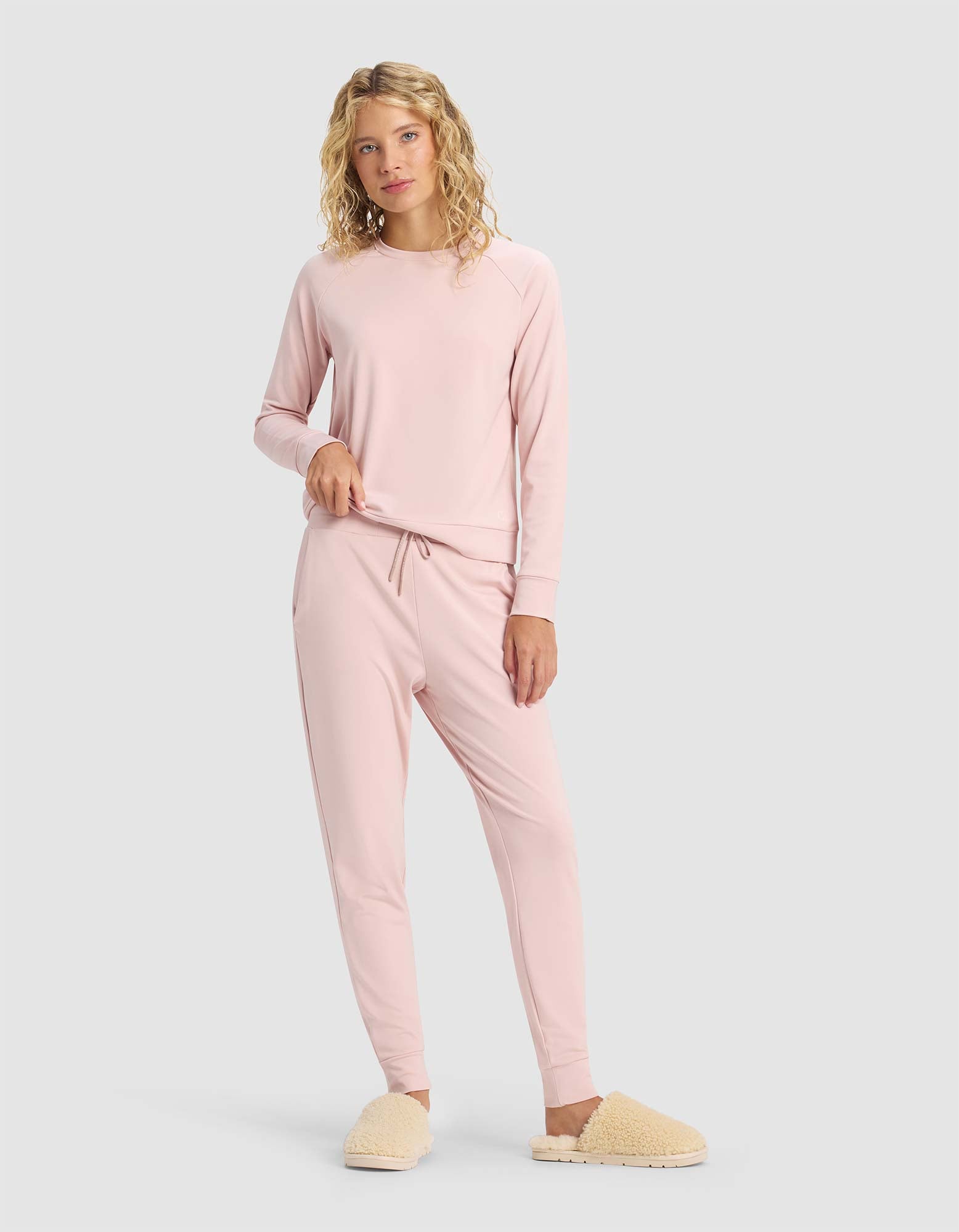 A person with curly blonde hair models the Cozy Earth Women's Brushed Bamboo Jogger Pant in light pink, paired with a matching long sleeve top and beige fuzzy slippers, standing against a plain light gray background.