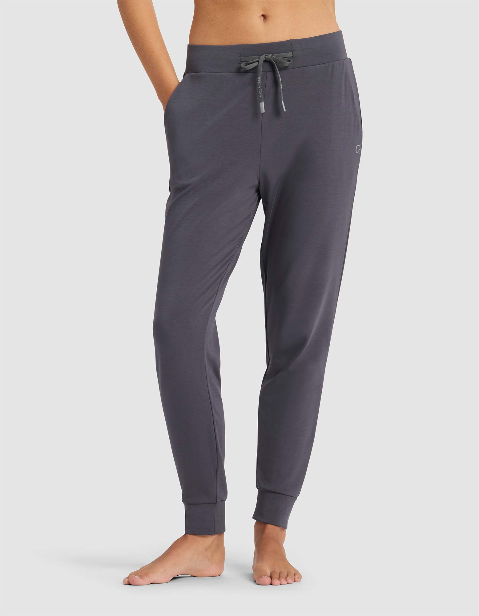 A person models Cozy Earth's Women's Brushed Bamboo Jogger Pant in dark gray, featuring a drawstring waist and pockets, standing barefoot against a plain light background. Upper body is not shown.