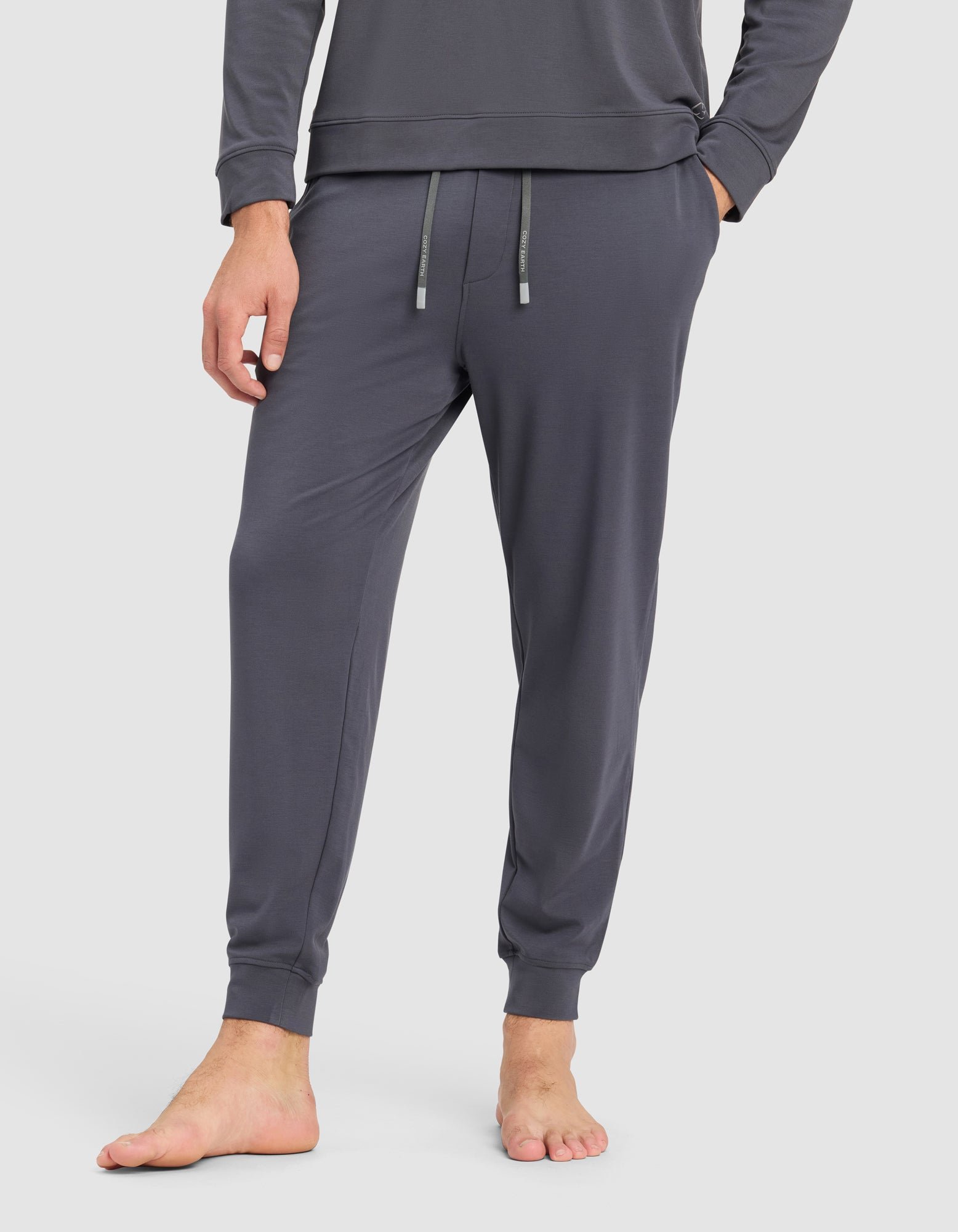 A person stands barefoot, showing only the lower half, wearing Cozy Earth's Men's Brushed Bamboo Jogger Pant in dark gray with an elastic waistband and drawstring.|Color:Graphite
