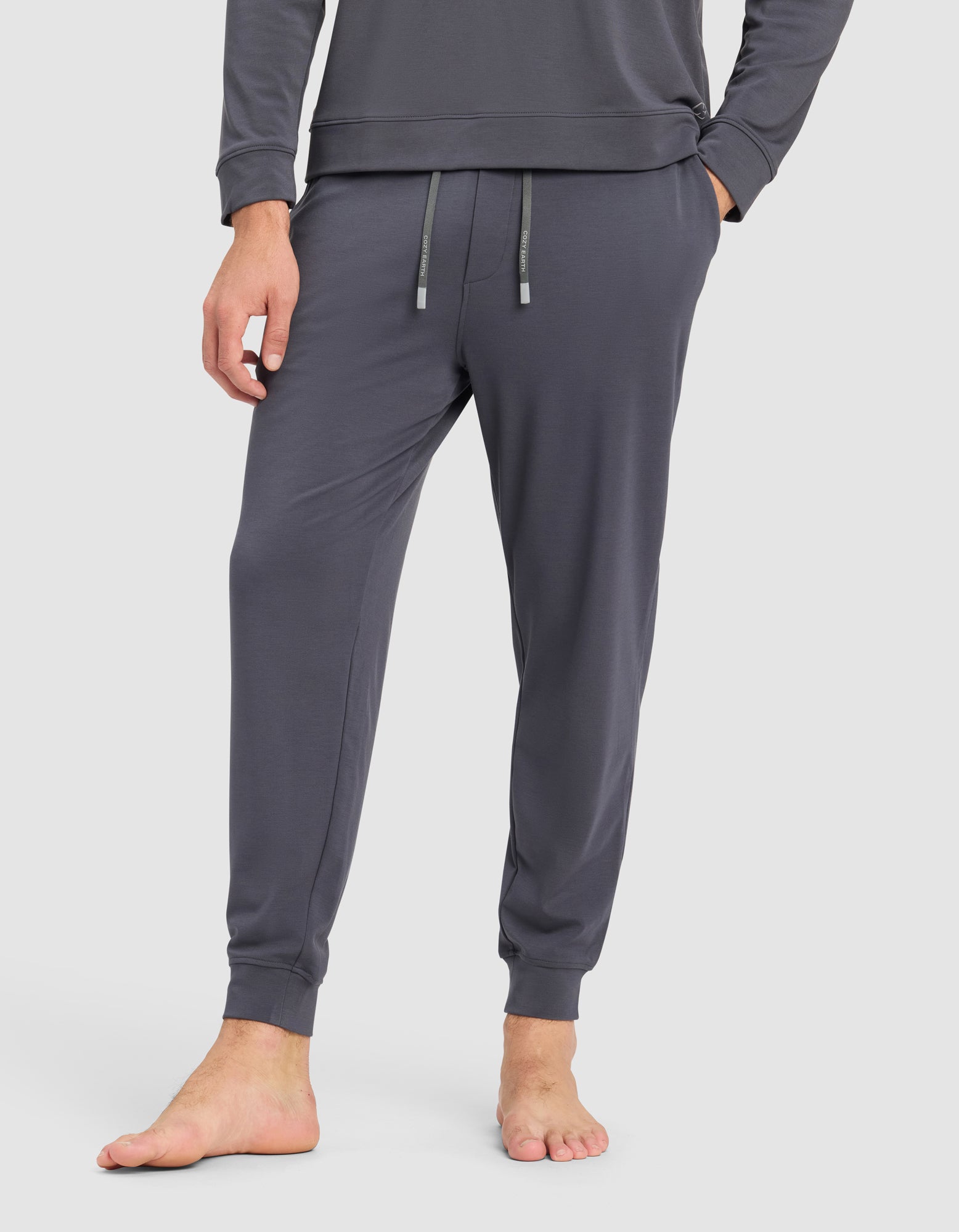 A person stands barefoot in Cozy Earth’s Men's Brushed Bamboo Hoodie & Jogger Set, featuring dark gray joggers with a drawstring waist and a matching long-sleeve hoodie, against a plain white background.
