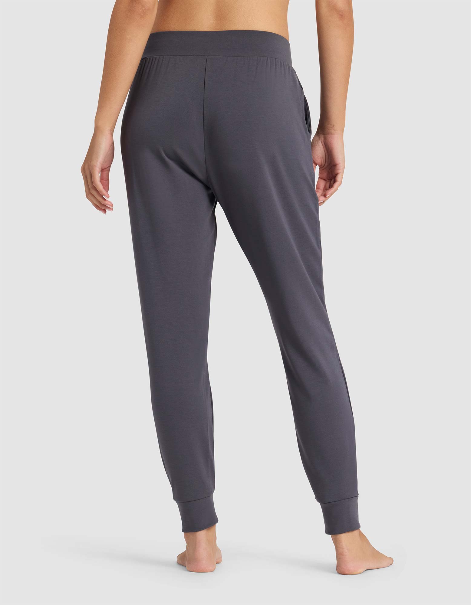 A person stands barefoot facing away, wearing Cozy Earth's Women's Brushed Bamboo Jogger Pant in dark gray with an elastic waistband and cuffed ankles, set against a plain light gray background.