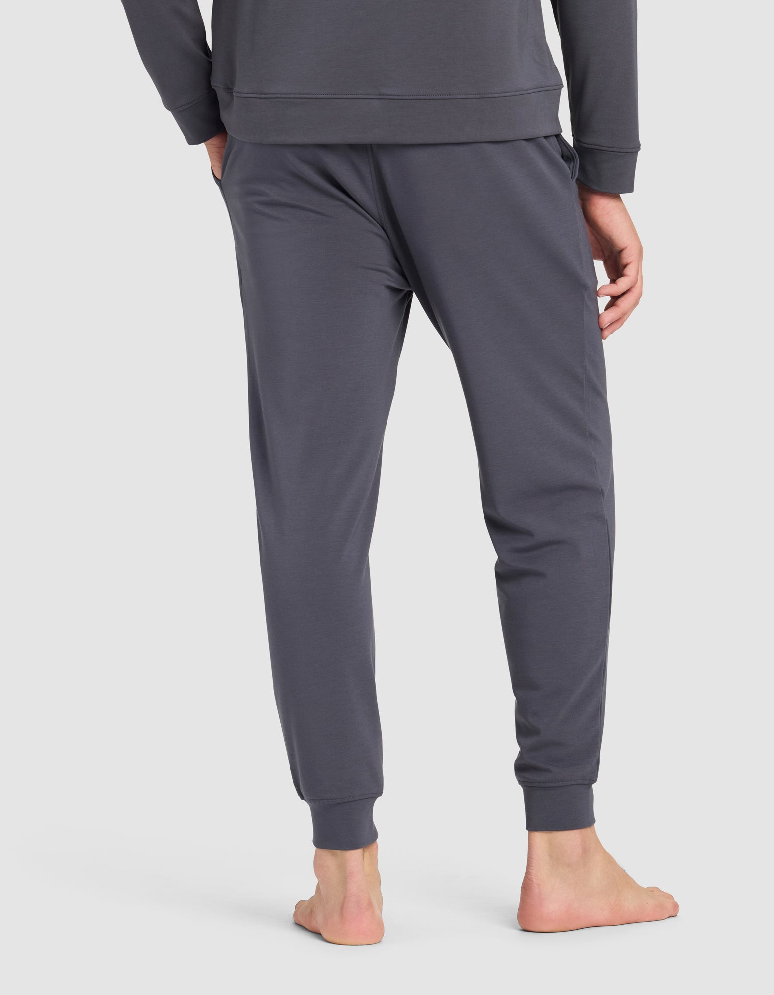 A person stands barefoot, back facing the camera, wearing Cozy Earth's Men's Brushed Bamboo Hoodie & Jogger Set in dark gray against a plain light background.