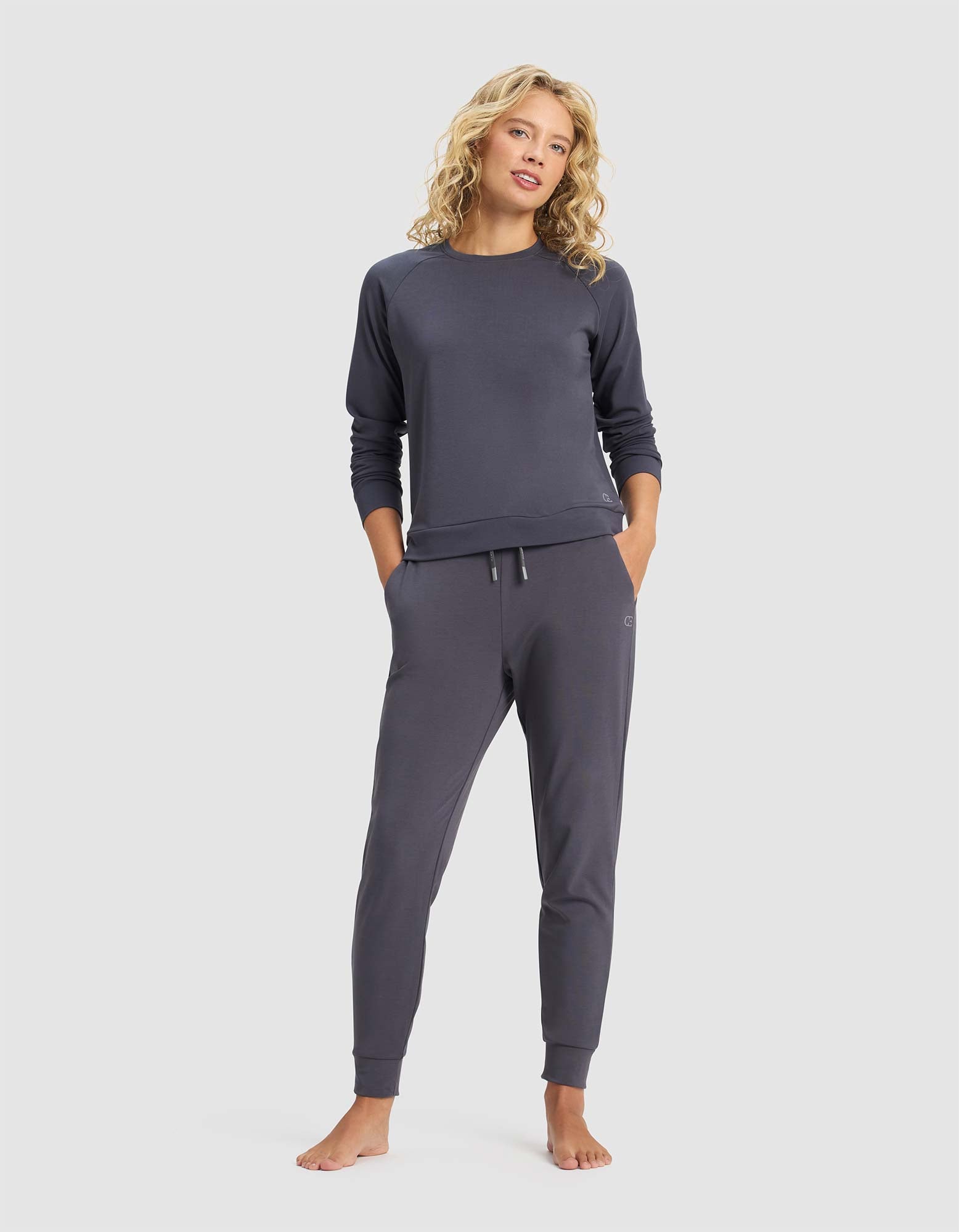A woman with curly blonde hair stands barefoot against a plain light background, wearing Cozy Earth's Women's Brushed Bamboo Jogger Pant and a matching dark gray long-sleeve top, with her hands in her pockets.