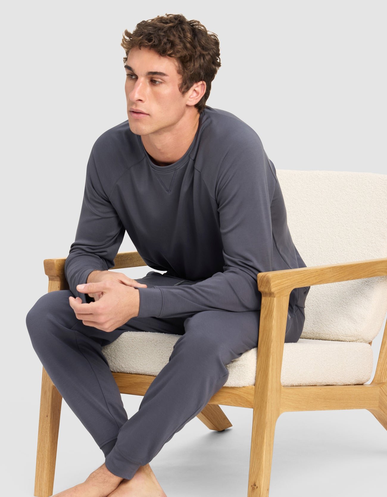 A young man sits barefoot on a cushioned wooden chair, wearing Cozy Earth's Men's Brushed Bamboo Jogger Pant in gray with a matching long-sleeve top. He looks forward with clasped hands against a plain, light gray background.