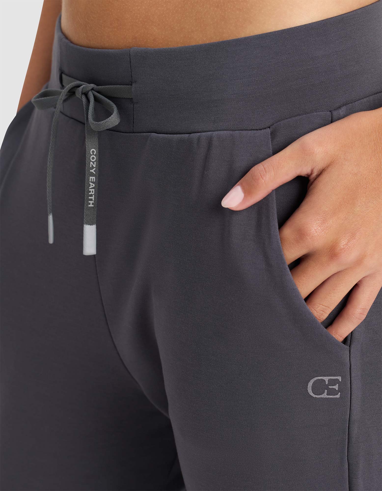 A woman wears Cozy Earth's Women's Brushed Bamboo Jogger Pant in dark gray, featuring "COZY EARTH" on the drawstring and a "CE" logo near the pocket, with one hand casually placed in the pocket.