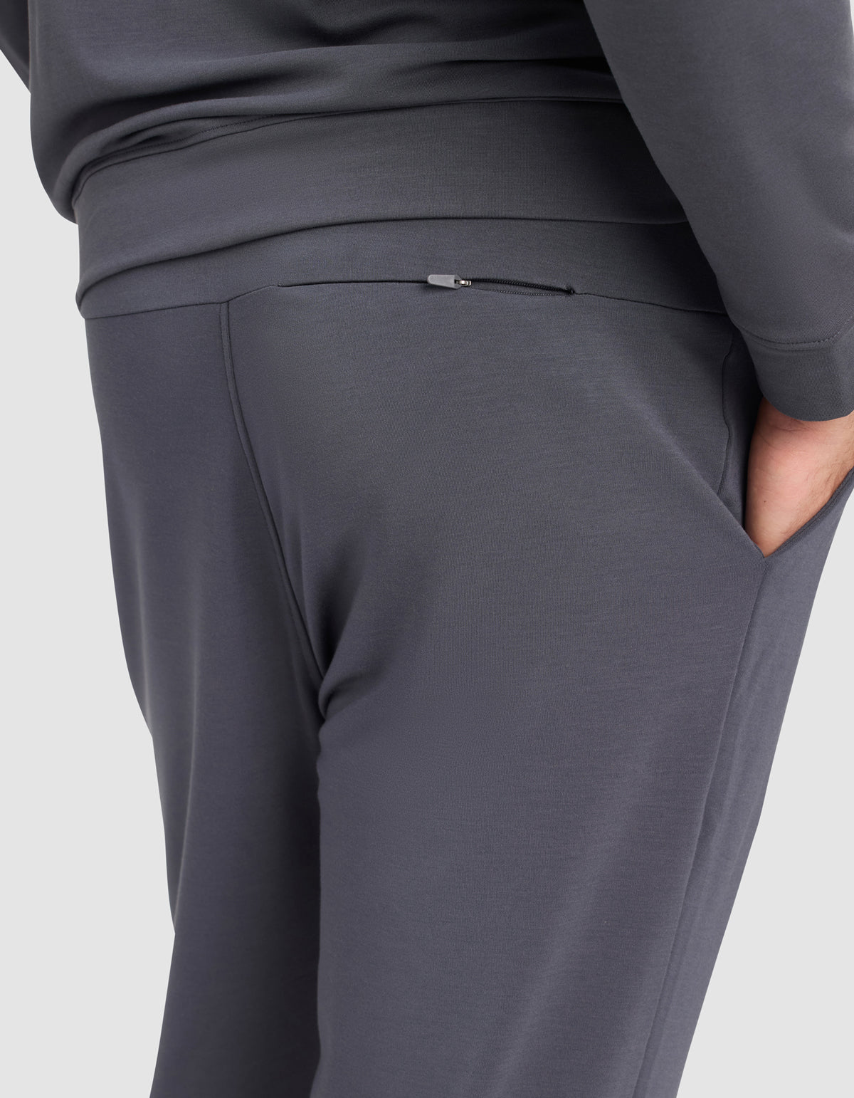 A person models the Cozy Earth Men's Brushed Bamboo Hoodie & Jogger Set in dark gray, standing sideways with one hand in their pocket to display the joggers' small zippered pocket detail at the back.
