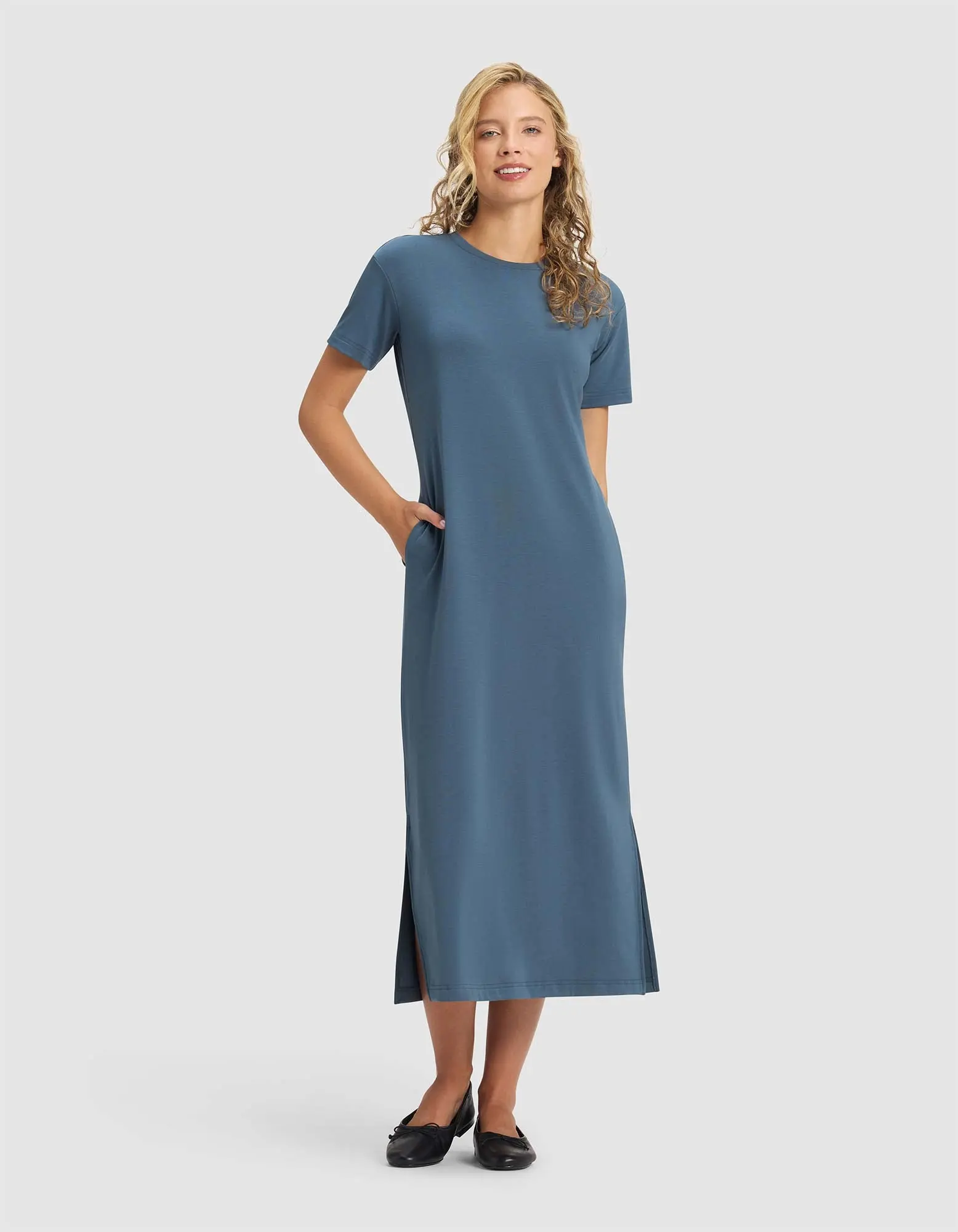 A woman smiles in the Cozy Earth Women's Brushed Bamboo Midi Dress, a blue, short-sleeve, ankle-length style with side slits and pockets. She pairs it with black flats. Her long blonde hair is loose against a plain light gray background. |Color:Dawn