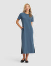 A woman smiles in the Cozy Earth Women's Brushed Bamboo Midi Dress, a blue, short-sleeve, ankle-length style with side slits and pockets. She pairs it with black flats. Her long blonde hair is loose against a plain light gray background. |Color:Dawn