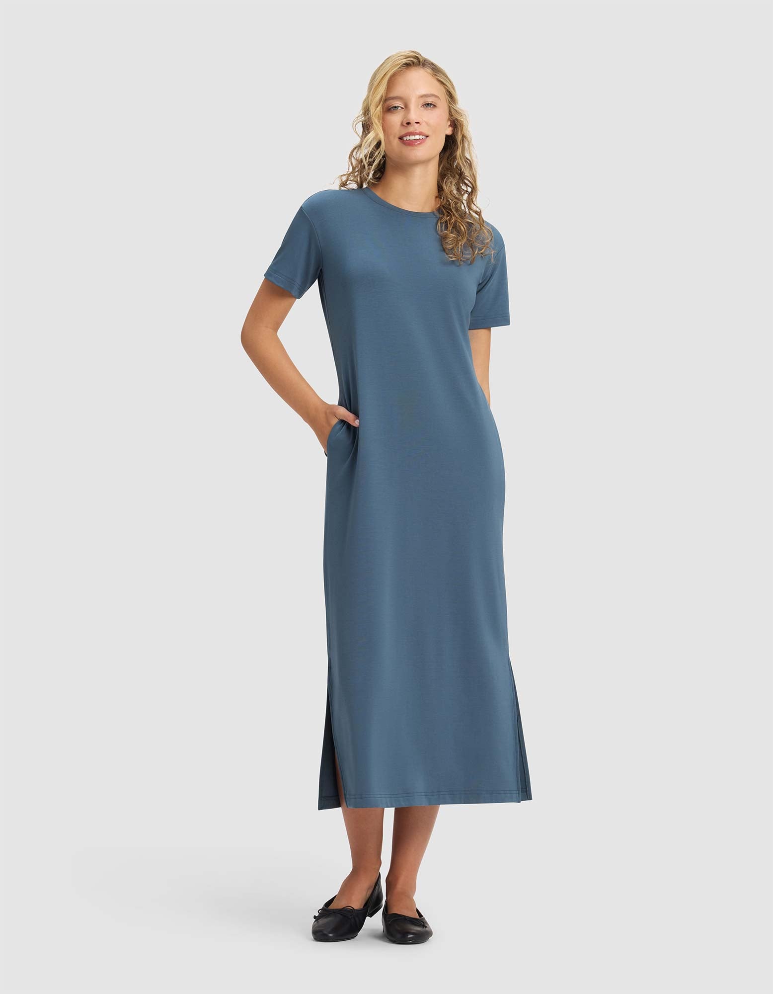 A woman smiles in the Cozy Earth Women's Brushed Bamboo Midi Dress, a blue, short-sleeve, ankle-length style with side slits and pockets. She pairs it with black flats. Her long blonde hair is loose against a plain light gray background. |Color:Dawn
