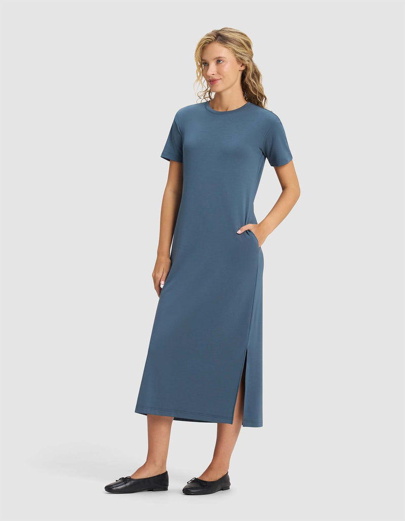 A woman wears the Cozy Earth Women's Brushed Bamboo Midi Dress—short-sleeve, ankle-length, blue with a side slit and pockets—standing against a plain light background and smiling slightly. She pairs it with black flats.