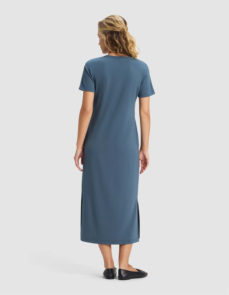 A woman with wavy blonde hair models Cozy Earth's Women's Brushed Bamboo Midi Dress, featuring short sleeves in teal-blue, standing in black shoes with her back to the camera against a light gray backdrop.