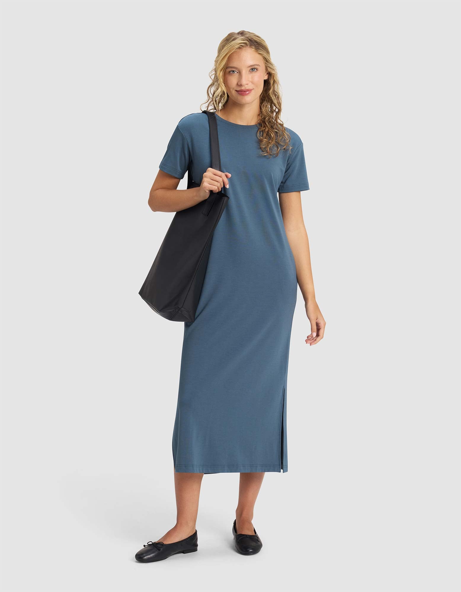 A woman with blonde, wavy hair wears the Cozy Earth Women's Brushed Bamboo Midi Dress in blue, featuring short sleeves and a side slit. She accessorizes with a large black tote and black loafers, standing against a plain light background. 