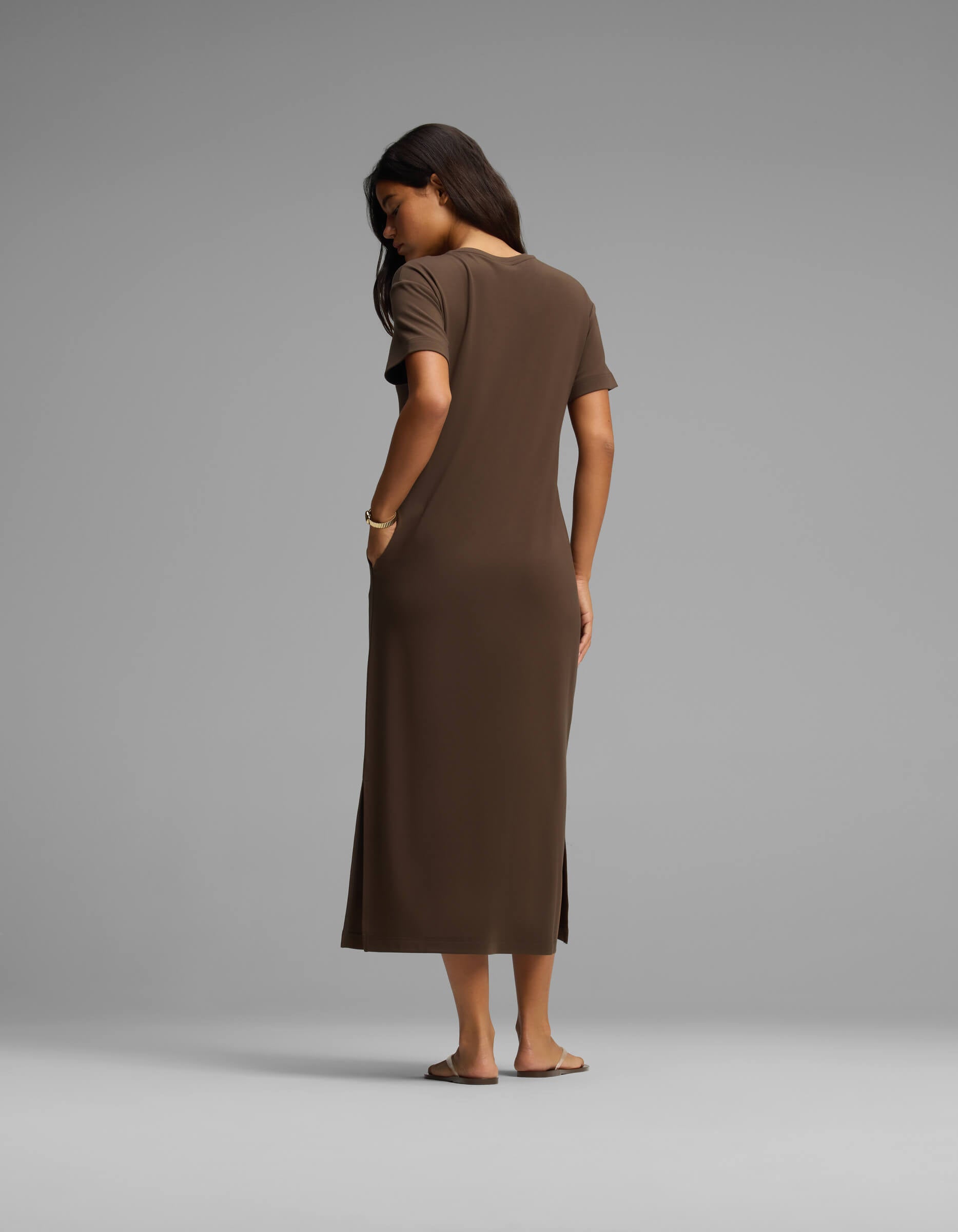 A woman stands barefoot facing away, wearing Cozy Earth's Women's Brushed Bamboo Midi Dress—a short-sleeve, ankle-length brown dress with her right hand in her pocket—set against a plain light gray background.|Color:Espresso